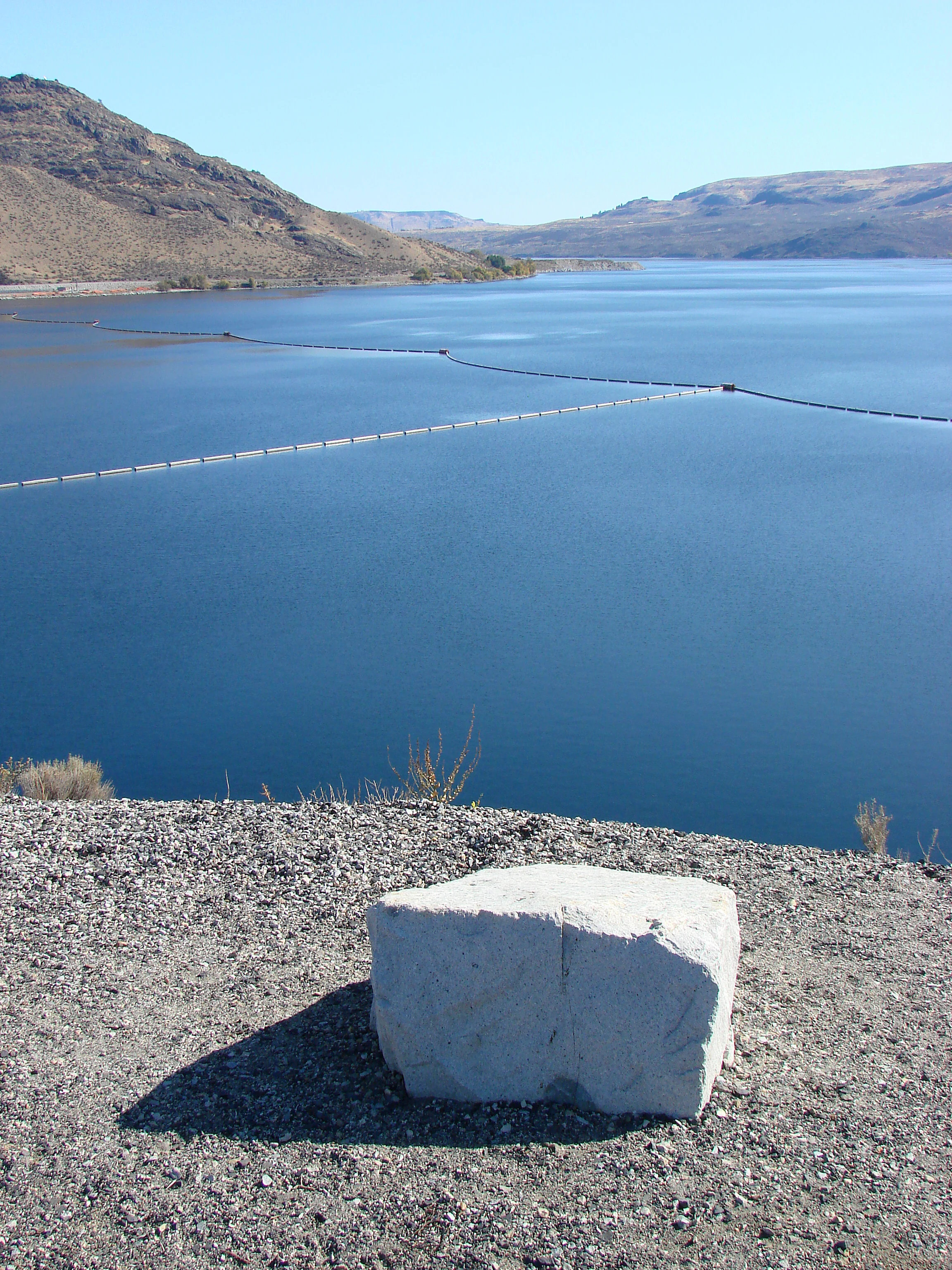 Una vista en ángulo alto de un embalse de aguas profundas y azules, con paisajes montañosos secos al fondo y una gran roca blanca en primer plano.