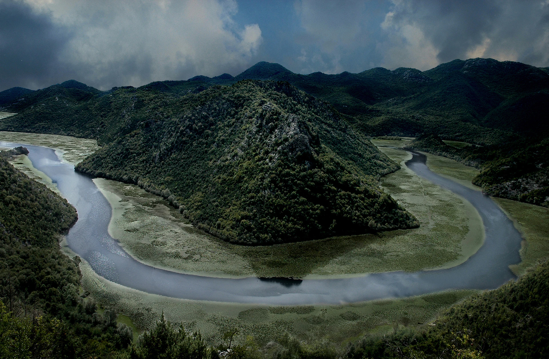 A high angle view of the Rijeka Crnojevica river looping in a dramatic horseshoe bend around a steep, green, forested hill, under a moody, partially cloudy sky.