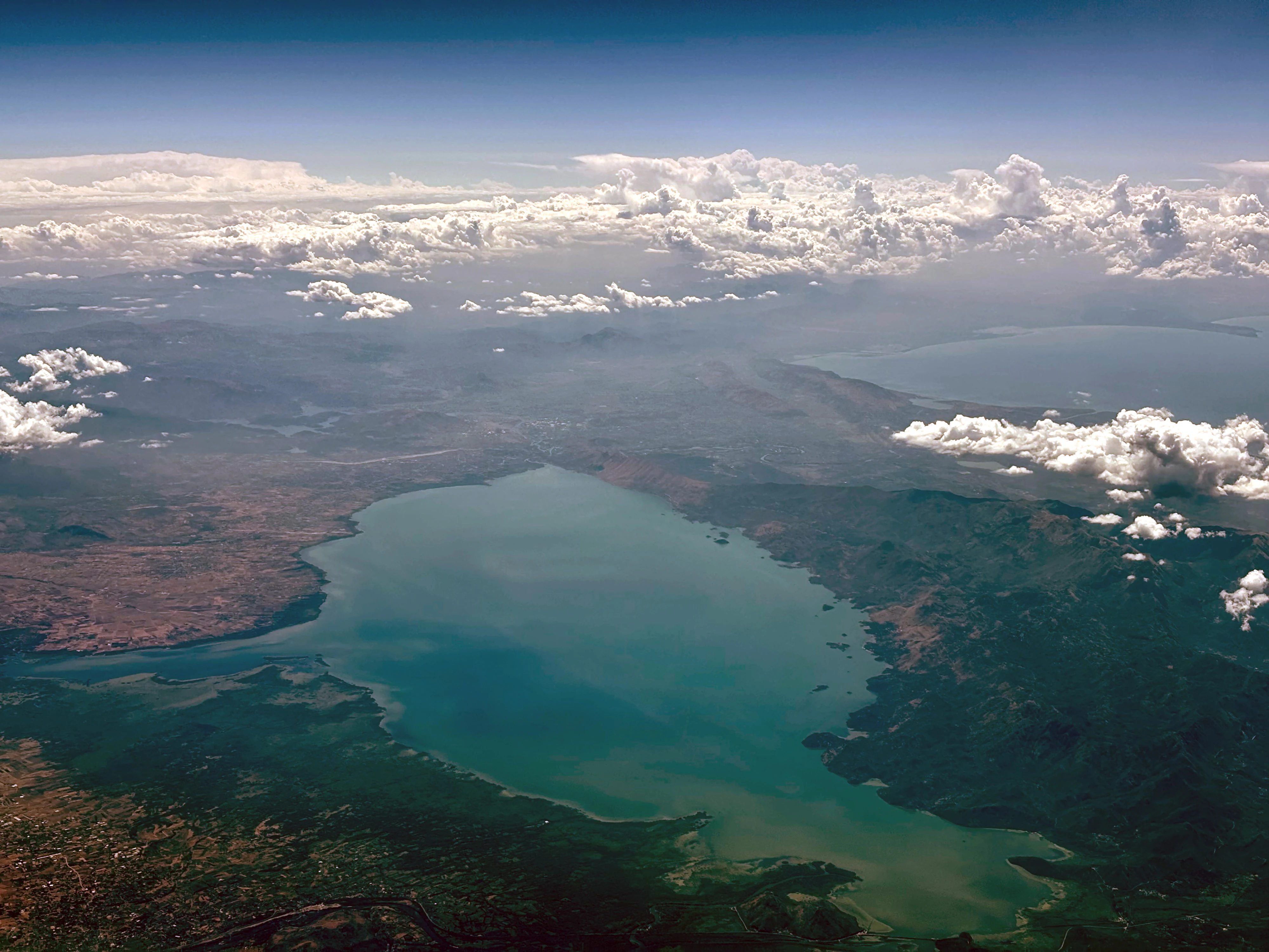 An aerial, wide-angle photograph looking down at Lake Skadar, featuring a large, blue-green lake basin surrounded by rolling hills, patches of dry vegetation, and scattered white clouds.