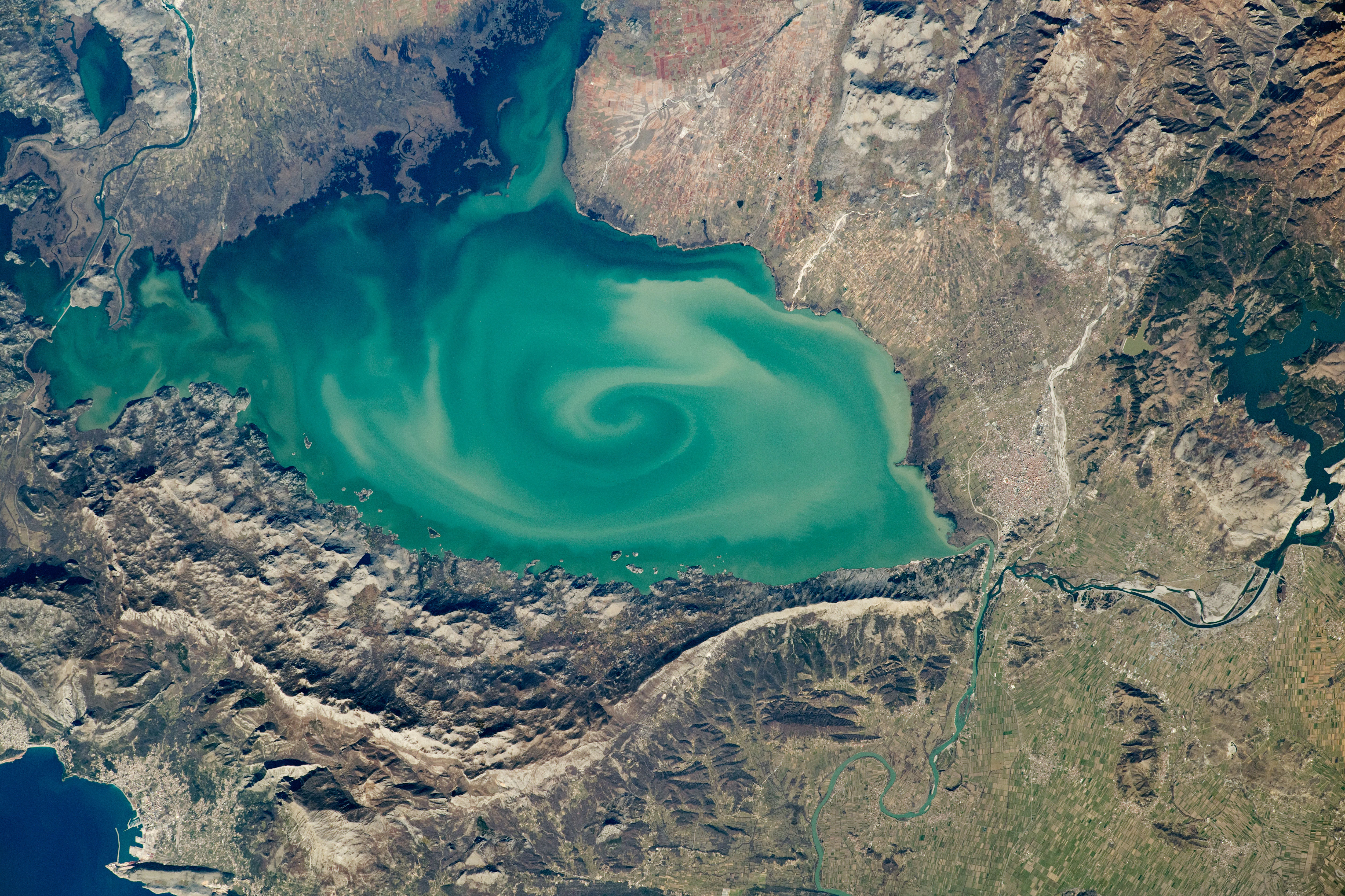Top-down satellite imagery of Lake Skadar, displaying a large body of turquoise water with visible swirling sediment patterns surrounded by rugged, mountainous terrain.