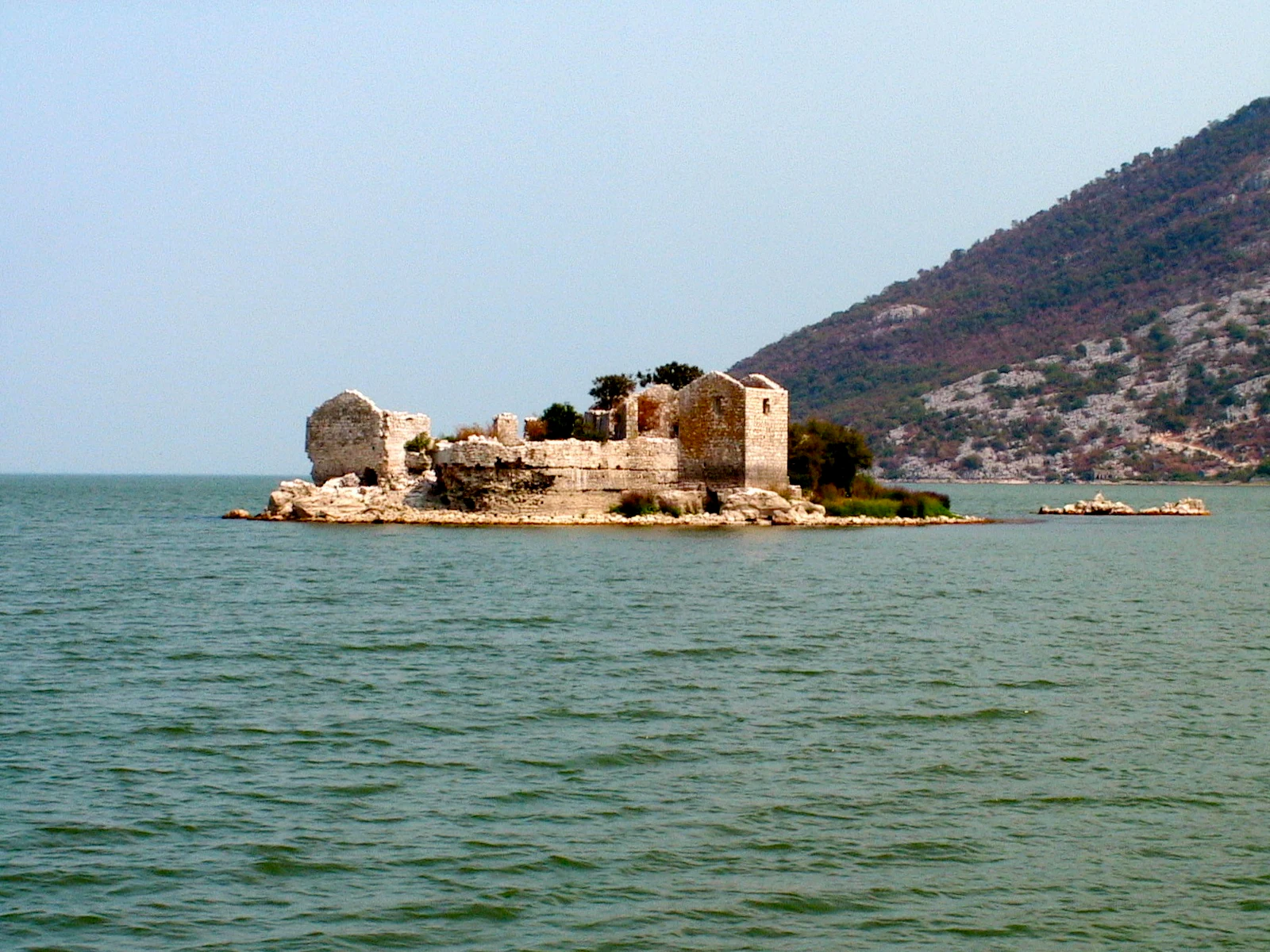 A small island with stone ruins of a fortress surrounded by lake water, with a large, rocky mountain slope in the background.