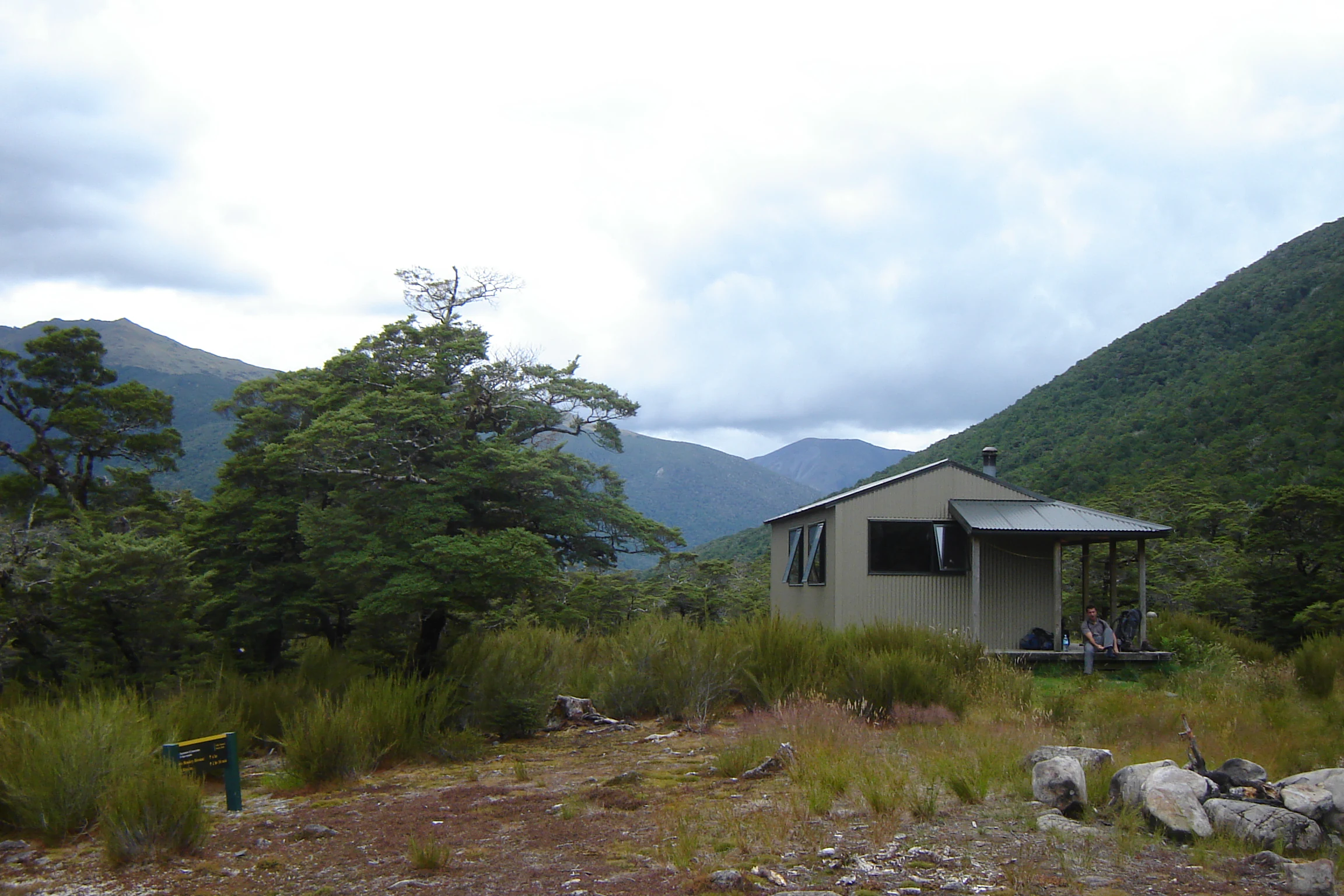 A small, light-colored backcountry hut with a porch nestled in a green valley surrounded by mountain slopes.