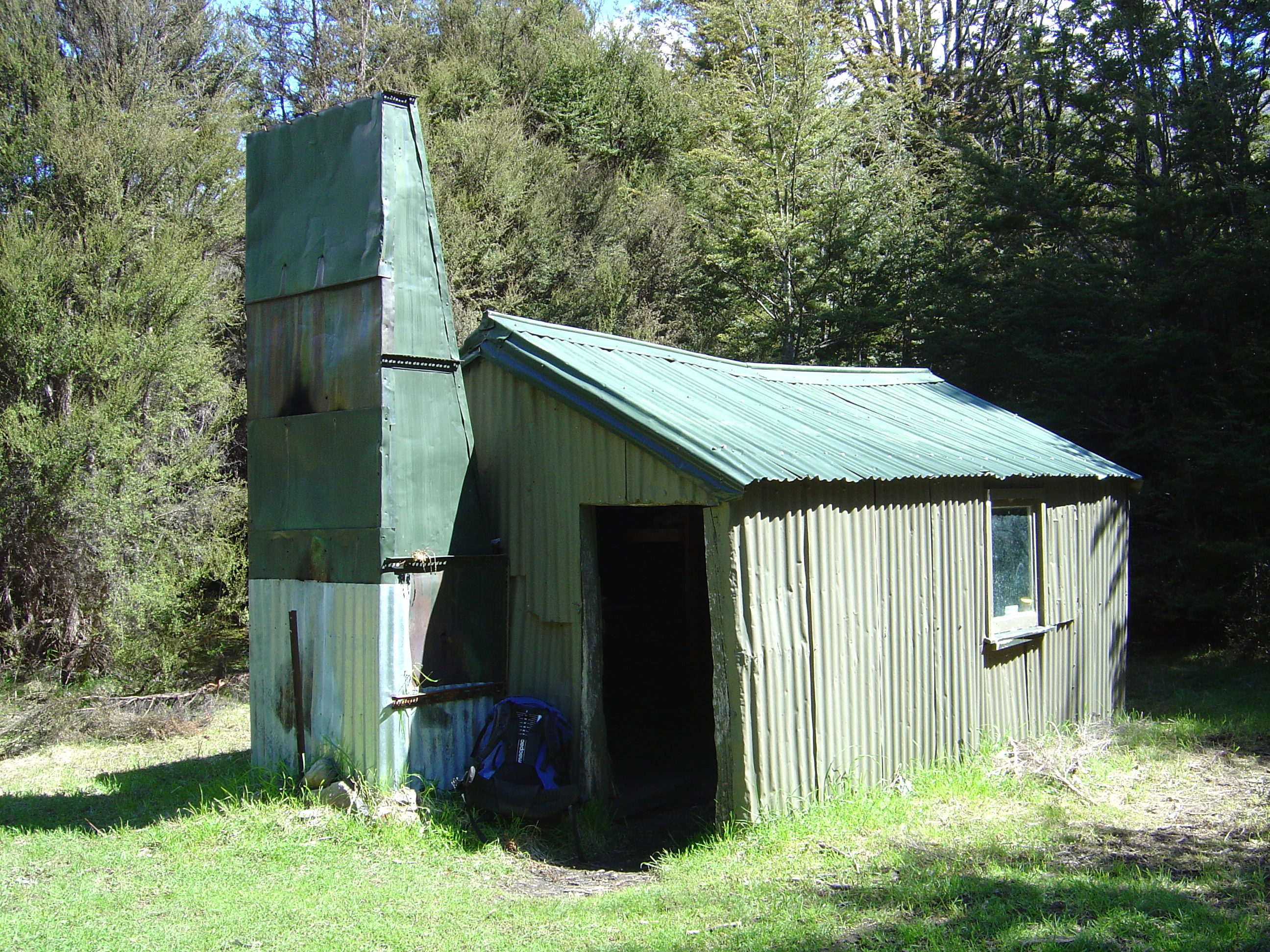 A small rustic backcountry hut with green corrugated metal walls and roof set against a background of dense forest trees.
