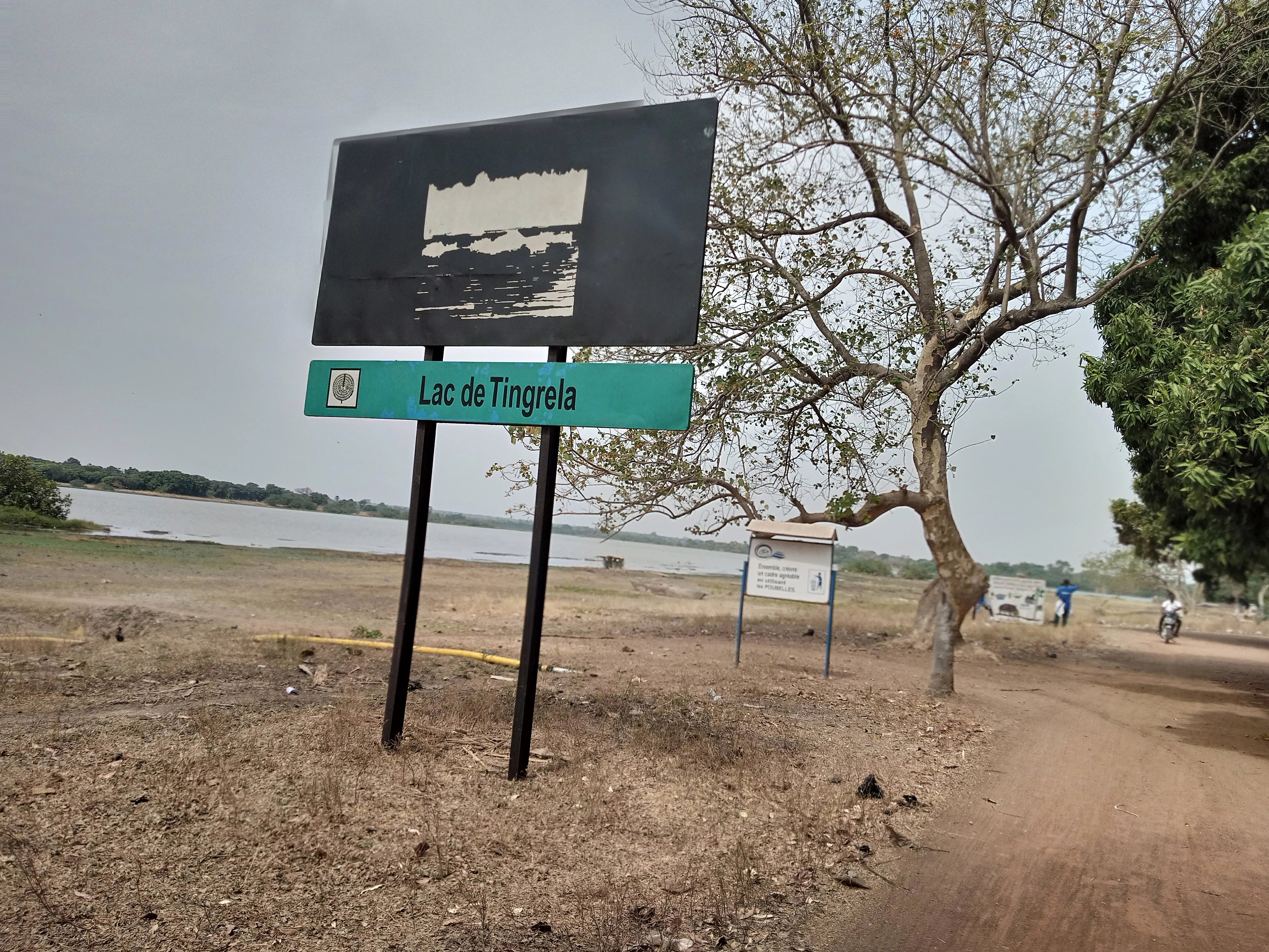 A metal sign with the text Lac de Tingrela mounted on two black poles, standing on a dirt road near a lake.