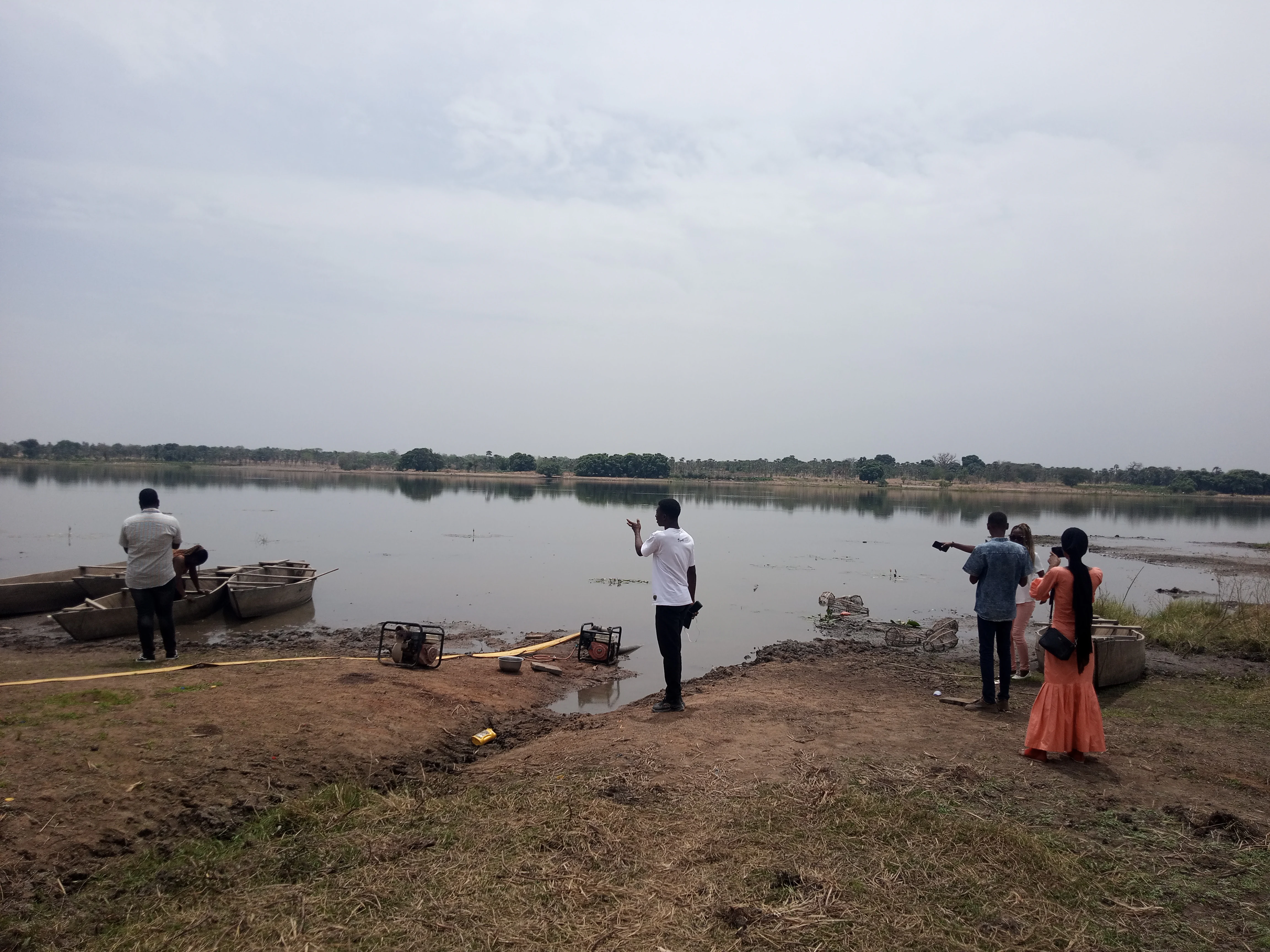 A group of people standing on the muddy shore of a calm lake, with several wooden boats moored at the water's edge under an overcast sky.