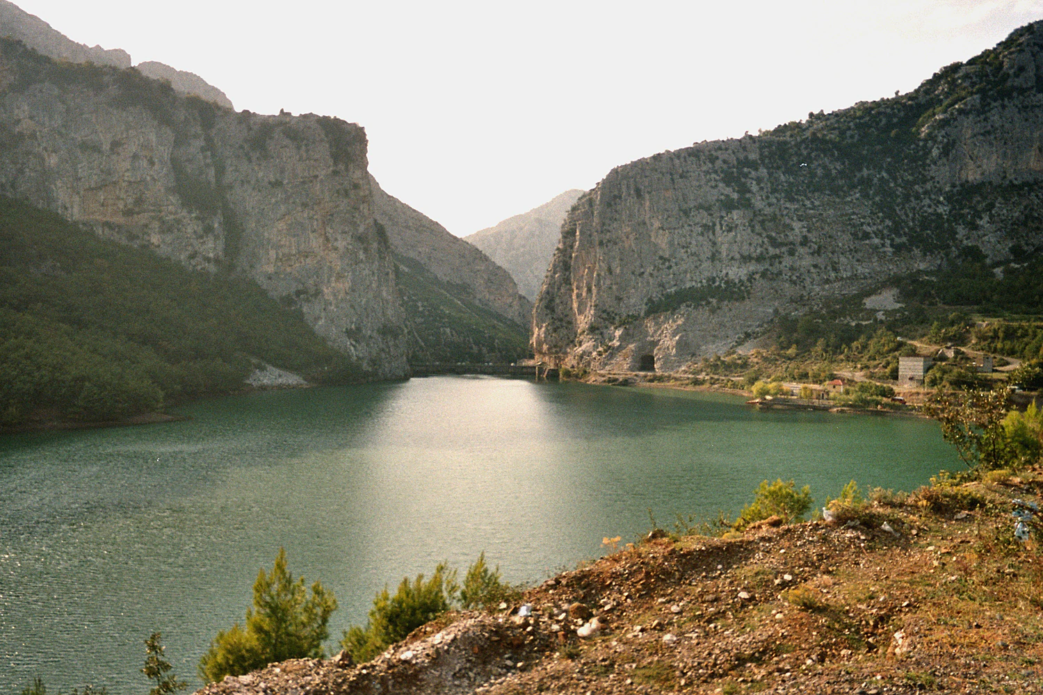 A wide, eye-level shot of a calm, turquoise lake nestled between two large, rocky, light-colored canyon walls, under a soft, hazy sky.