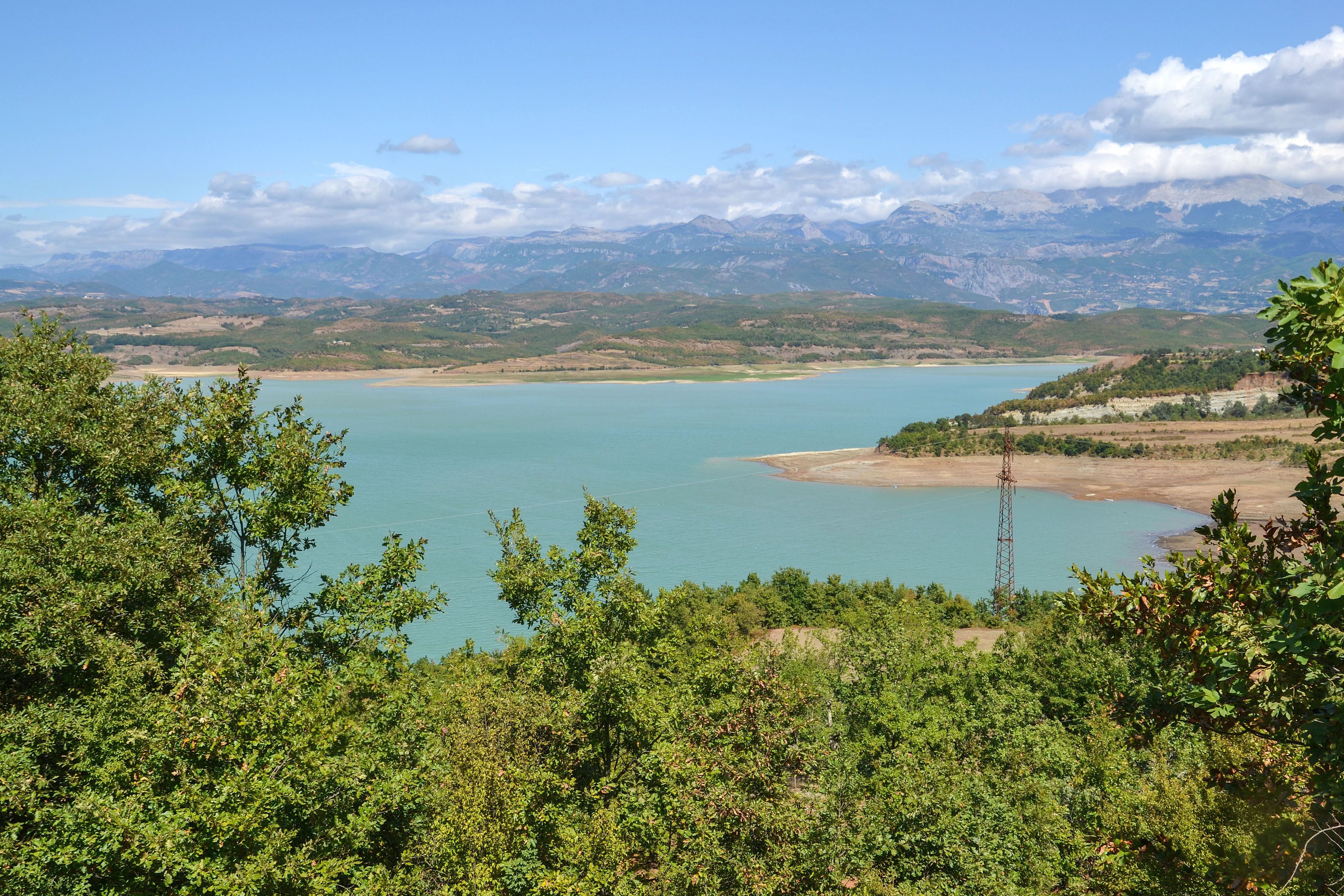 A wide shot overlooking the turquoise water of Lake Ulza, with dense green trees in the foreground, rolling hills in the midground, and distant mountains under a blue sky with clouds.