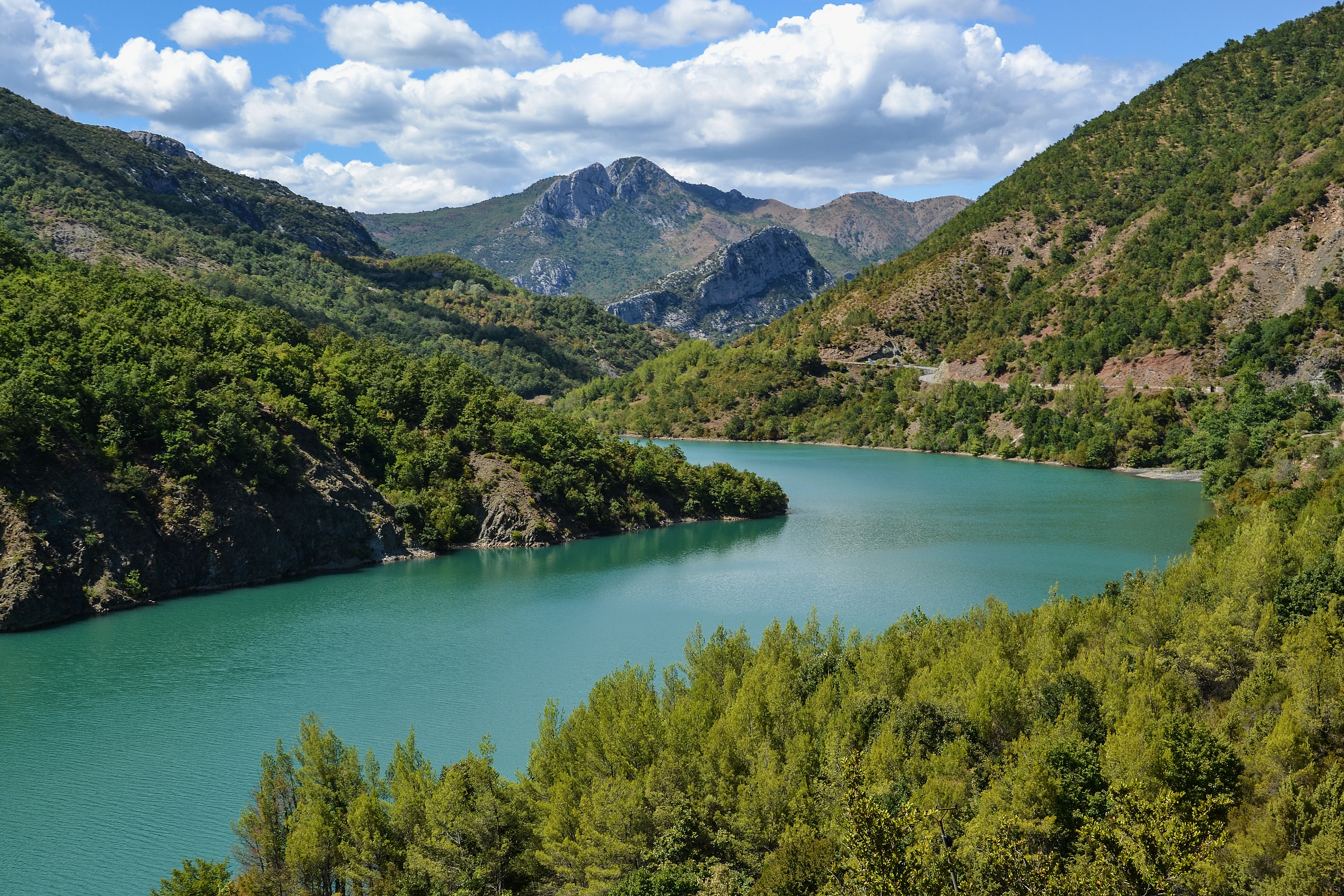 A wide-angle landscape shot of a turquoise river winding through a valley flanked by dense green forests and rugged mountains under a blue sky with white clouds.