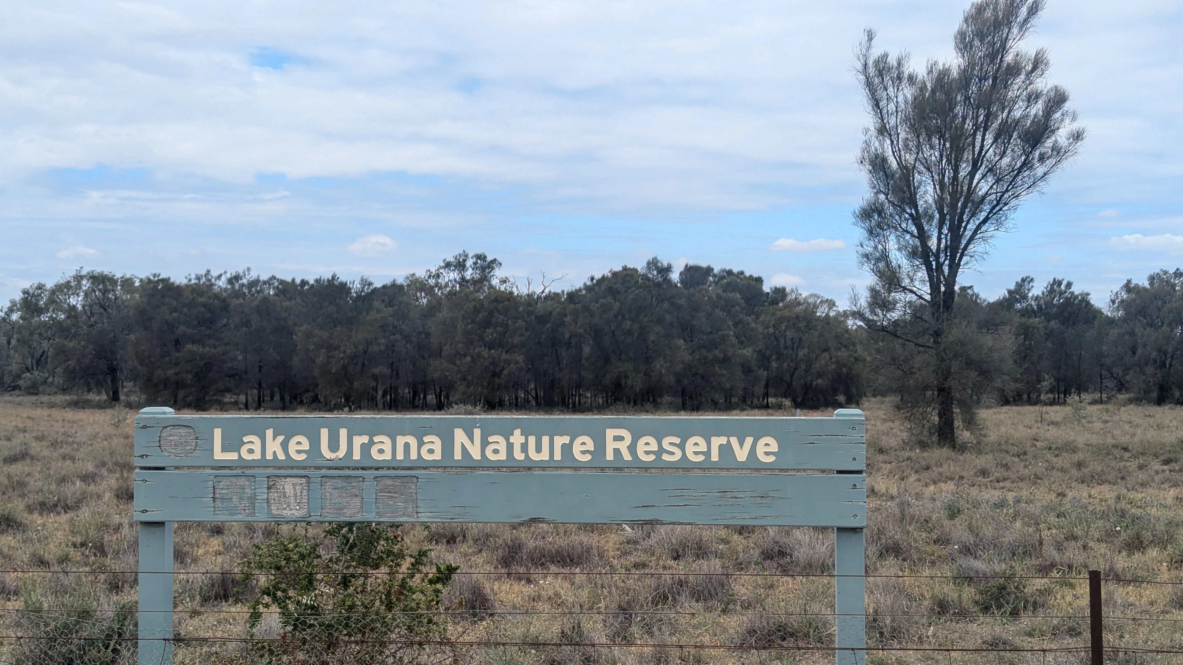 A pale green wooden park entrance sign reading Lake Urana Nature Reserve, set against a background of dry grasslands and scattered trees under a cloudy sky.