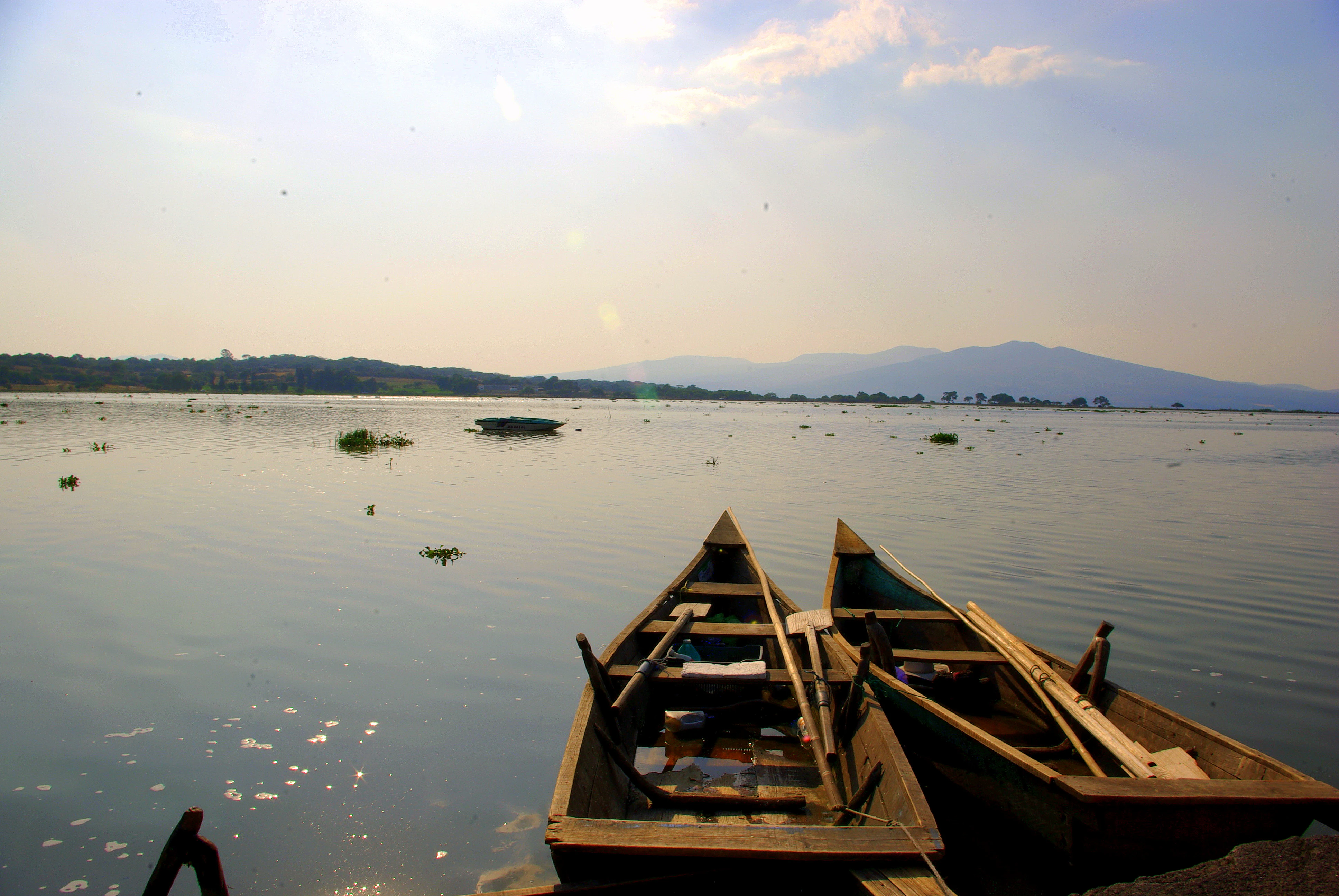 Two small wooden rowboats floating at the edge of a large, calm lake with distant hills visible under a hazy sky.