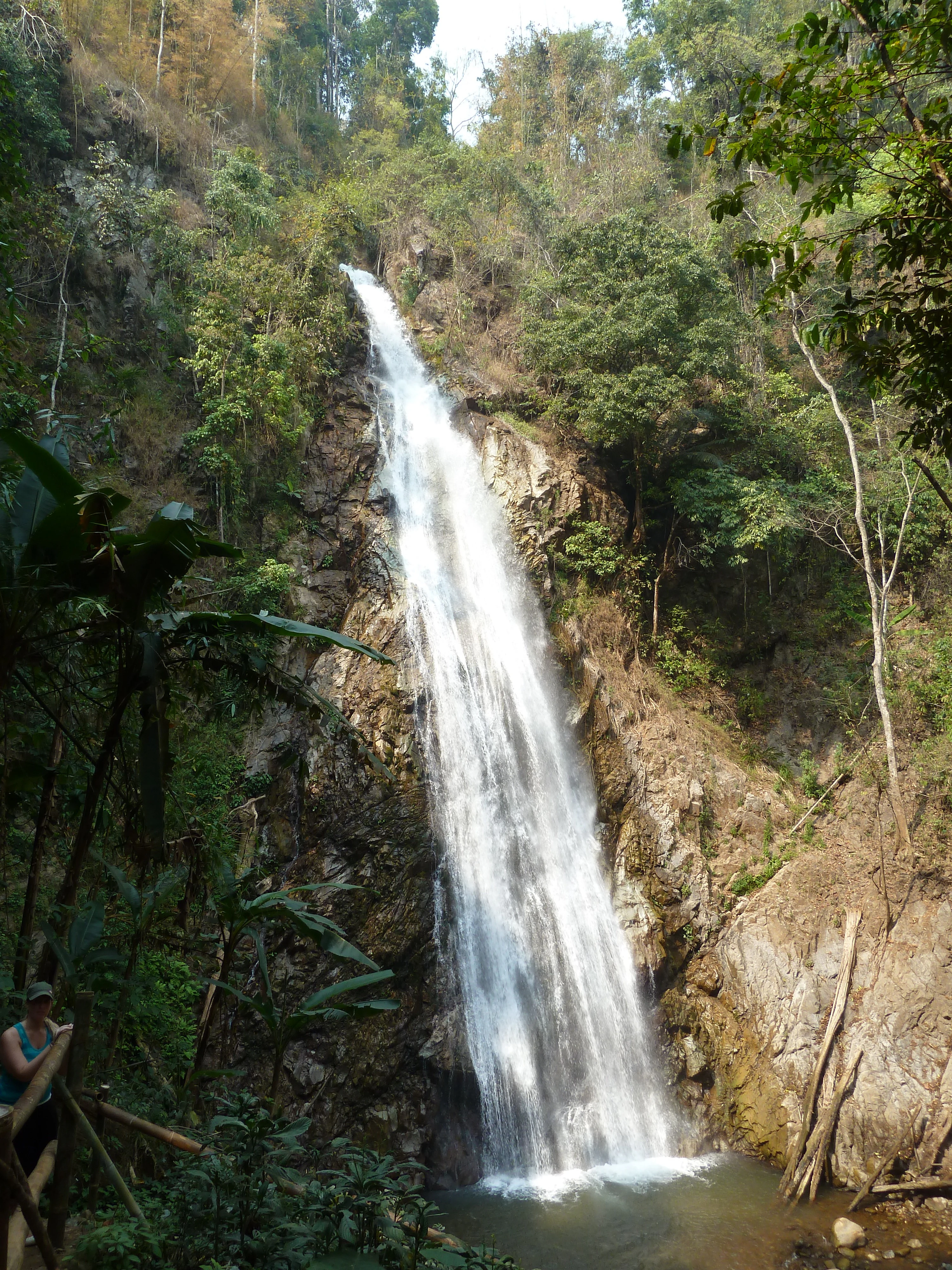 A tall waterfall cascading down a rocky cliff surrounded by dense green forest with a person holding a walking stick visible on the left