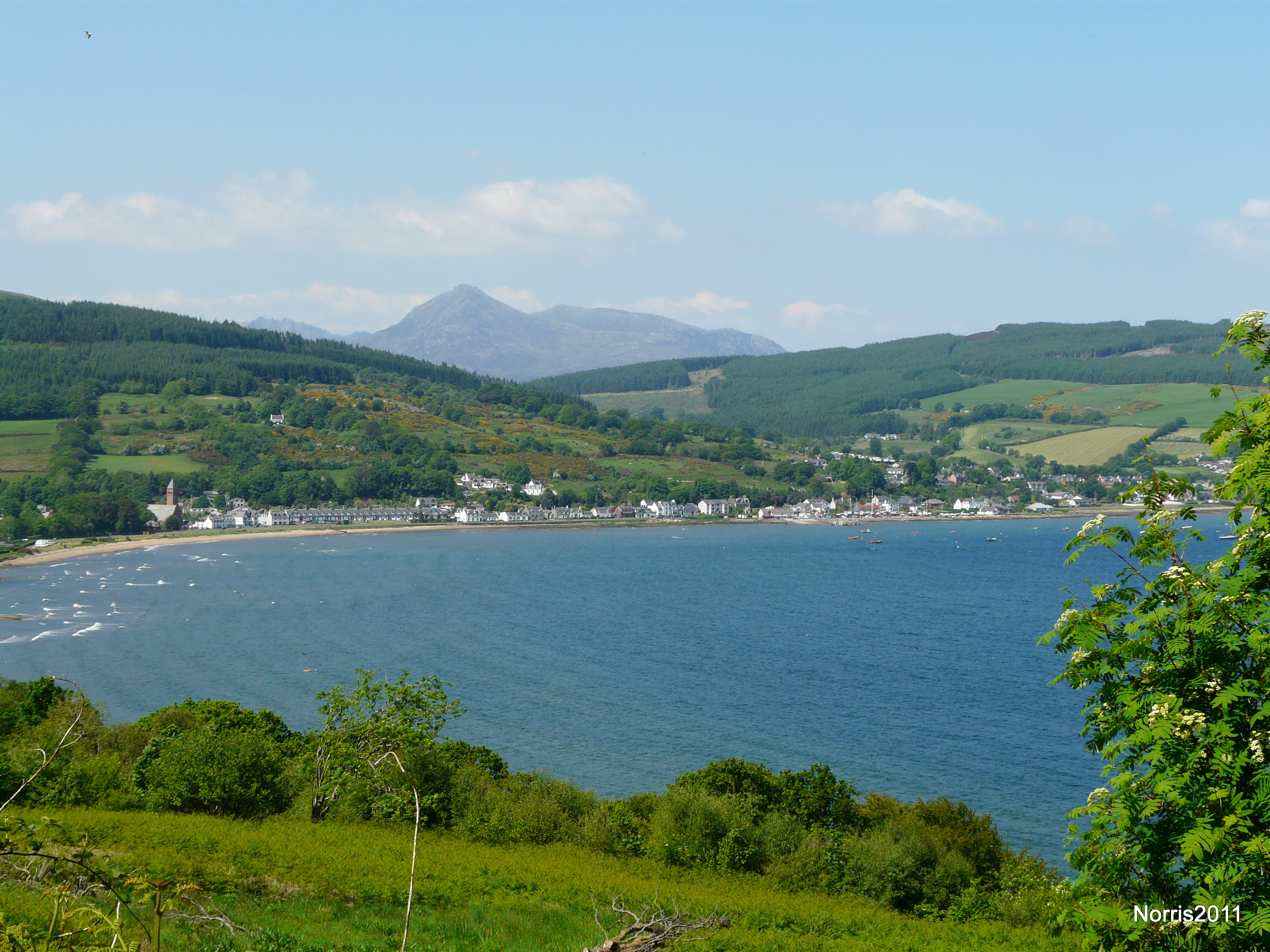 A wide shot looking across a blue bay towards a coastal village nestled between rolling green hills, with a mountain peak rising in the background under a clear sky.