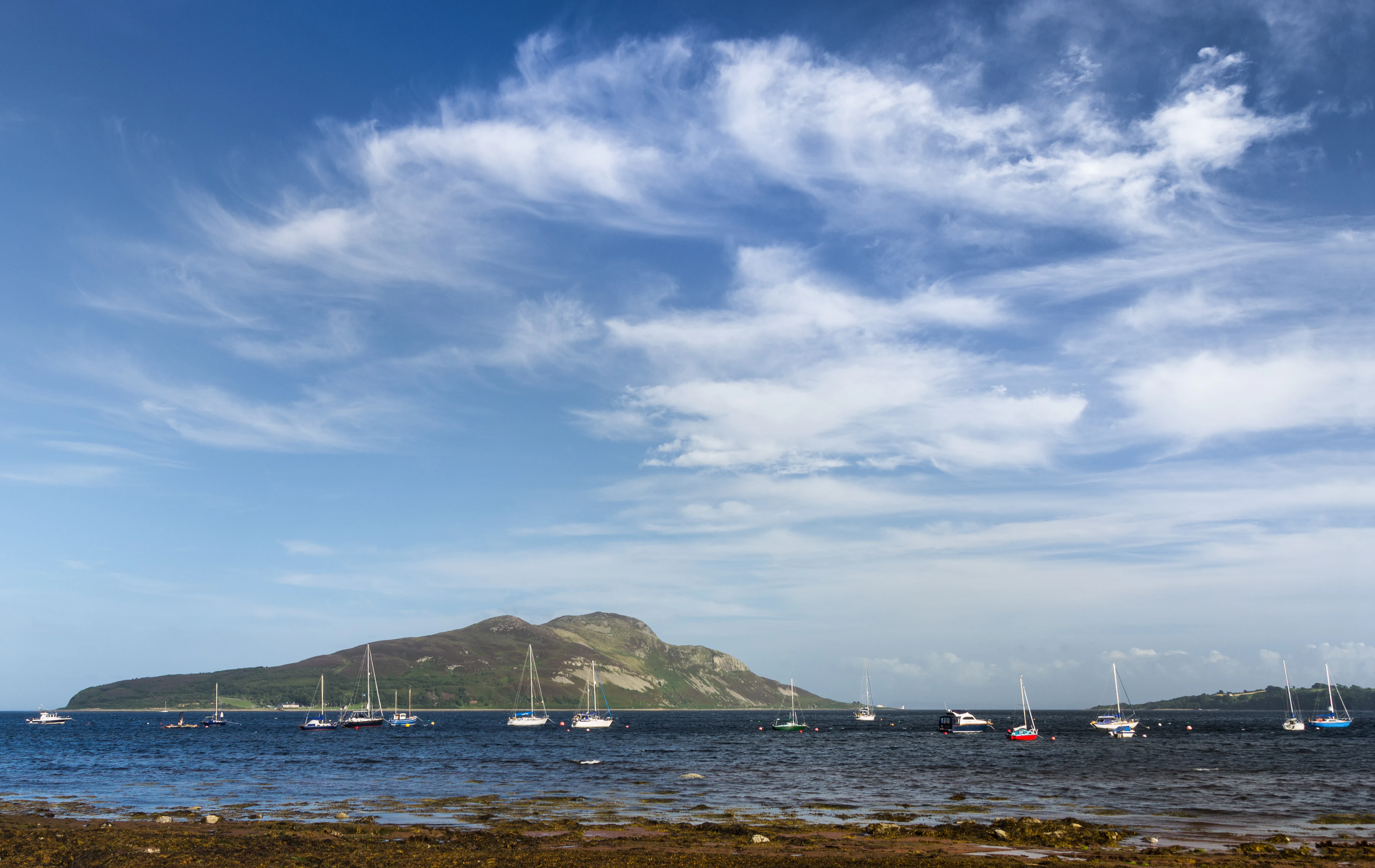 A wide view of a calm bay with numerous sailboats moored in the distance, framed by a large island hill and a bright blue sky with wispy clouds.
