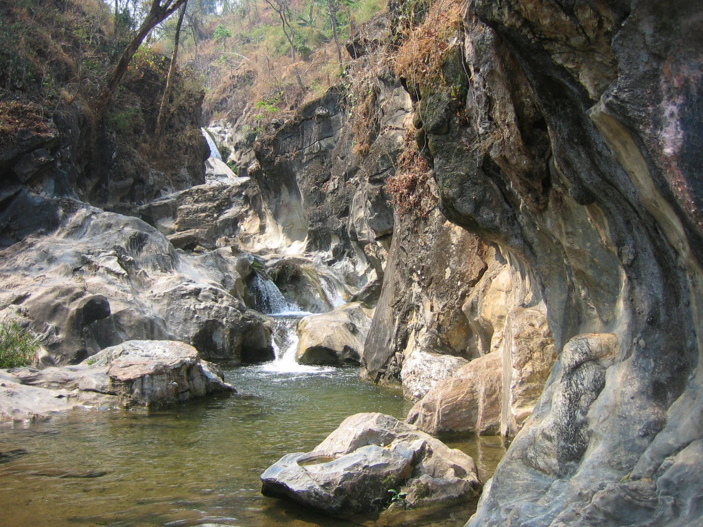 Cascada rocosa con un pequeño arroyo fluyendo sobre rocas hacia una poza, rodeada de vegetación y acantilados