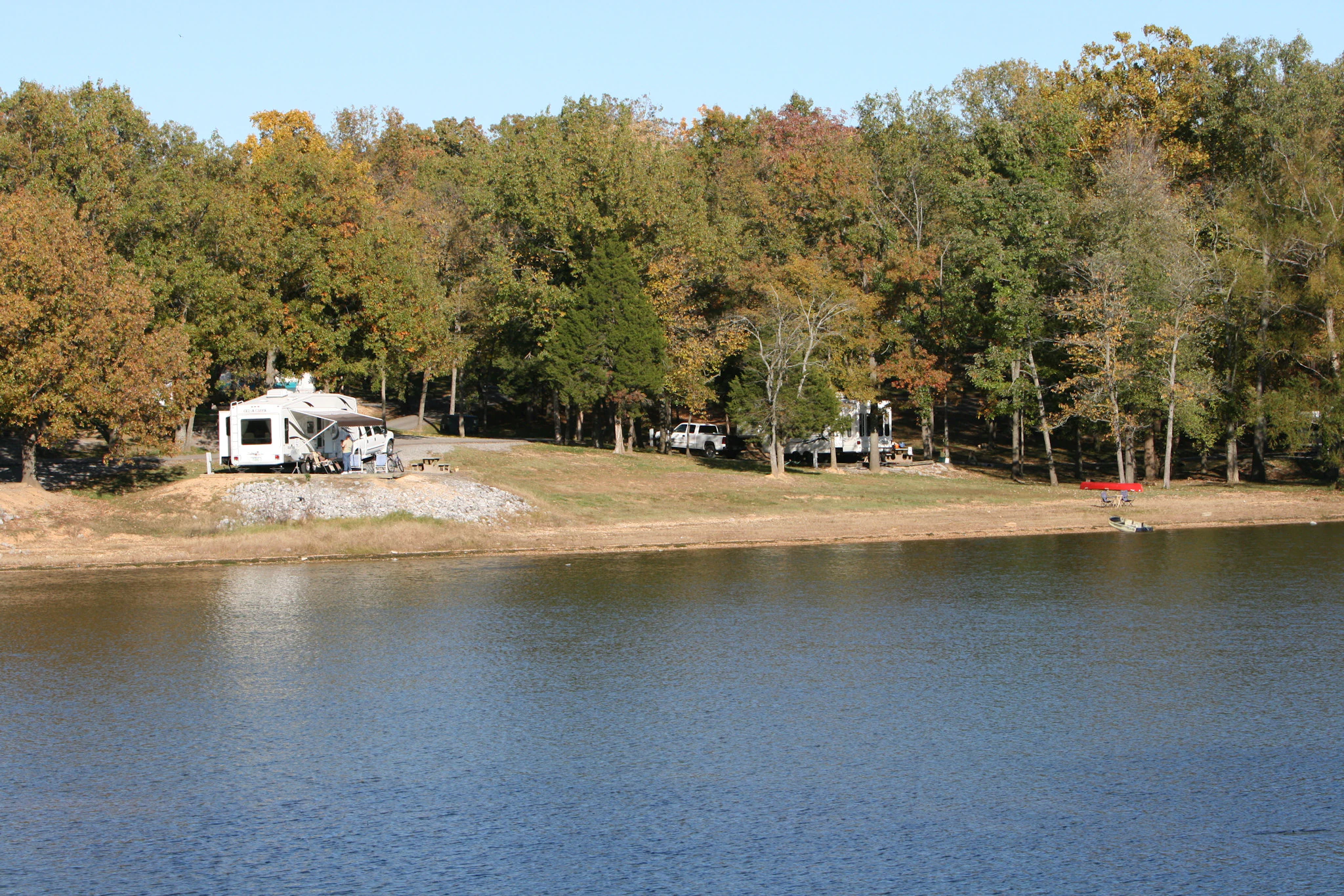 Varios vehículos recreativos estacionados en un campamento boscoso junto a un lago, a lo largo de una masa de agua tranquila durante la temporada de otoño.