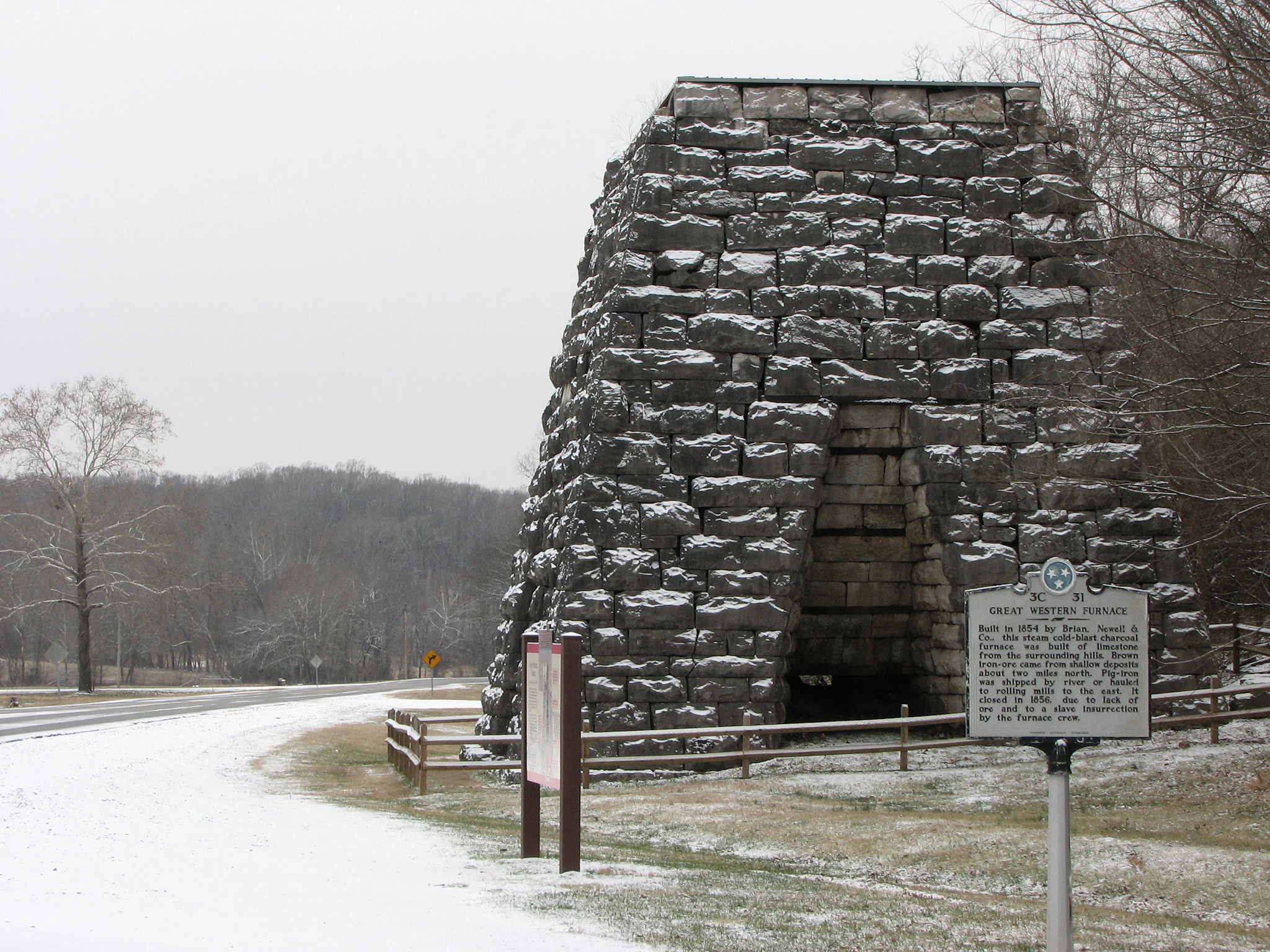 Una estructura de horno de hierro de piedra en invierno, con un diseño de bloque cónico y nieve en sus superficies, situada junto a un marcador histórico al borde de la carretera.