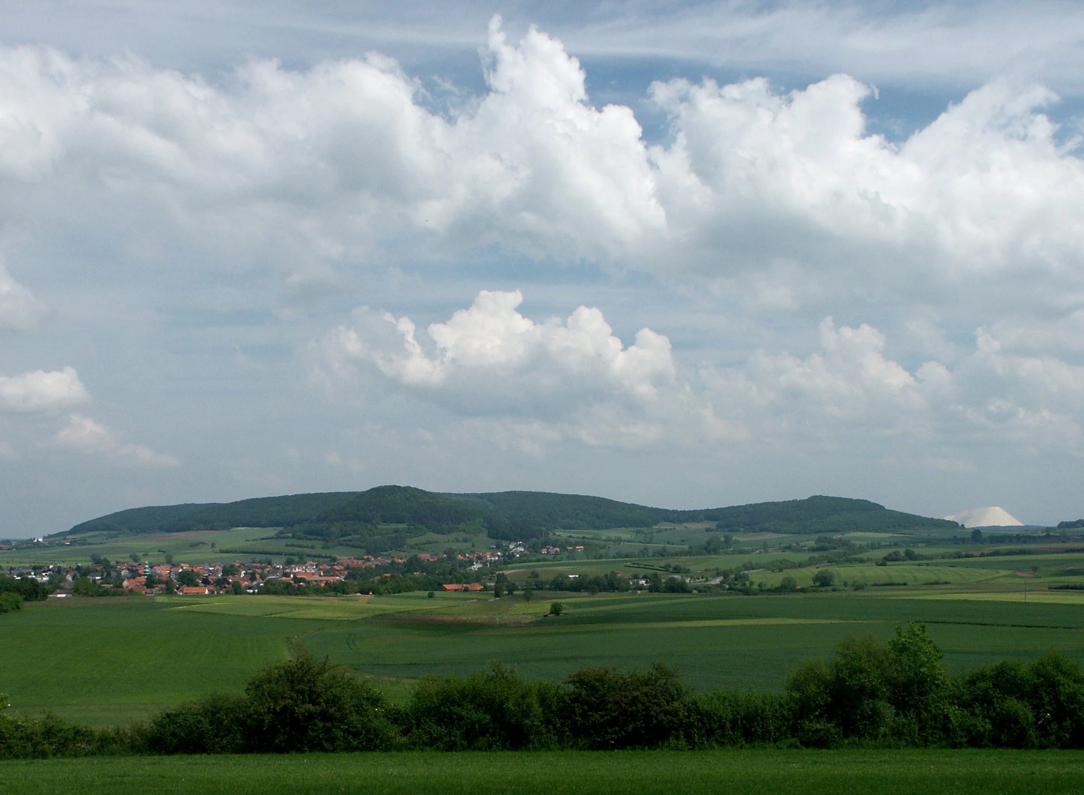 A wide-angle landscape photograph showing the gentle, forested slopes of Landecker Berg in the distance, framed by open green fields and a small village in the midground under a partly cloudy sky.