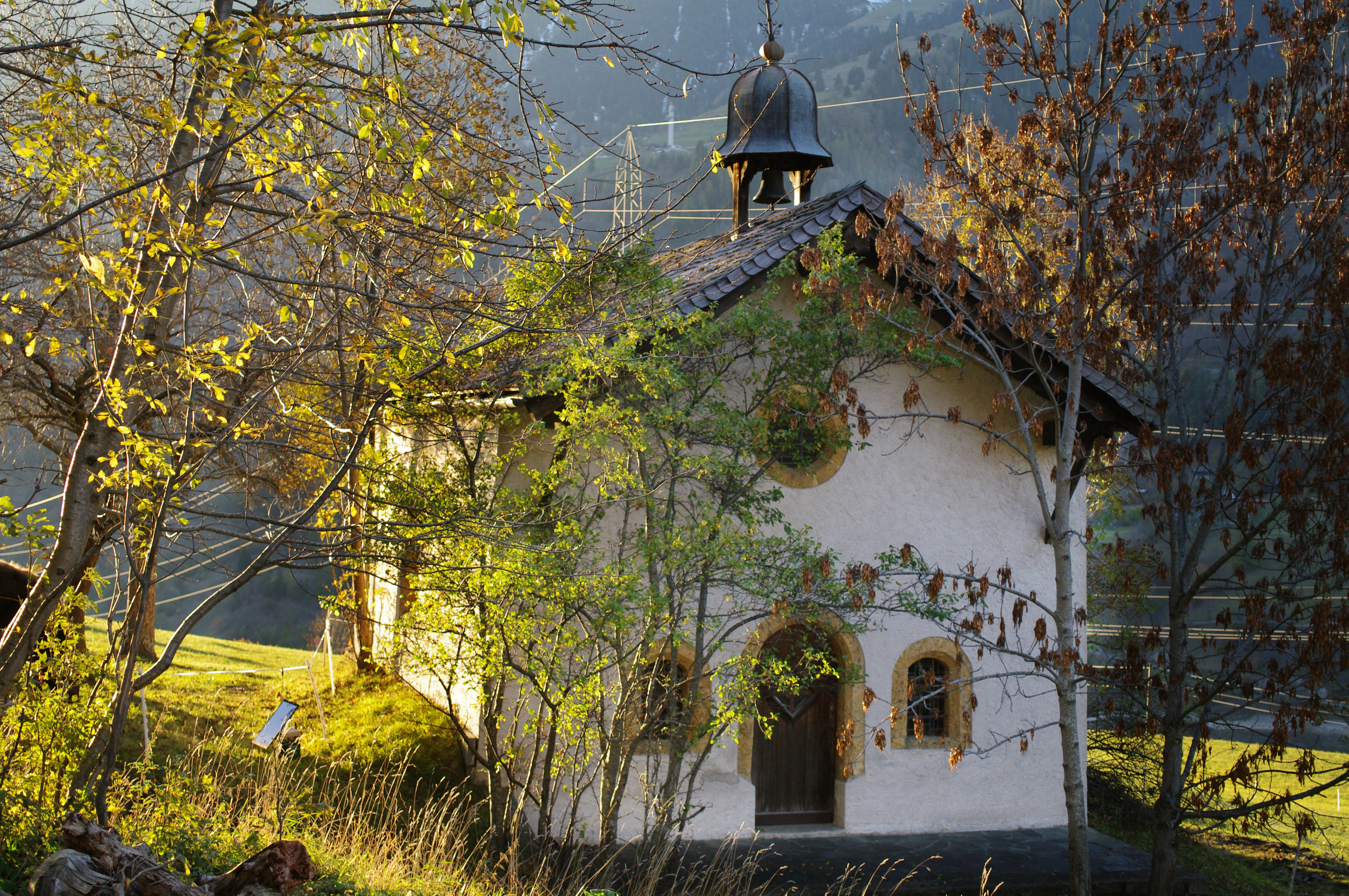 A small light-colored stone chapel with a dark bell tower, partially framed by trees with autumn foliage, situated in a mountain setting.