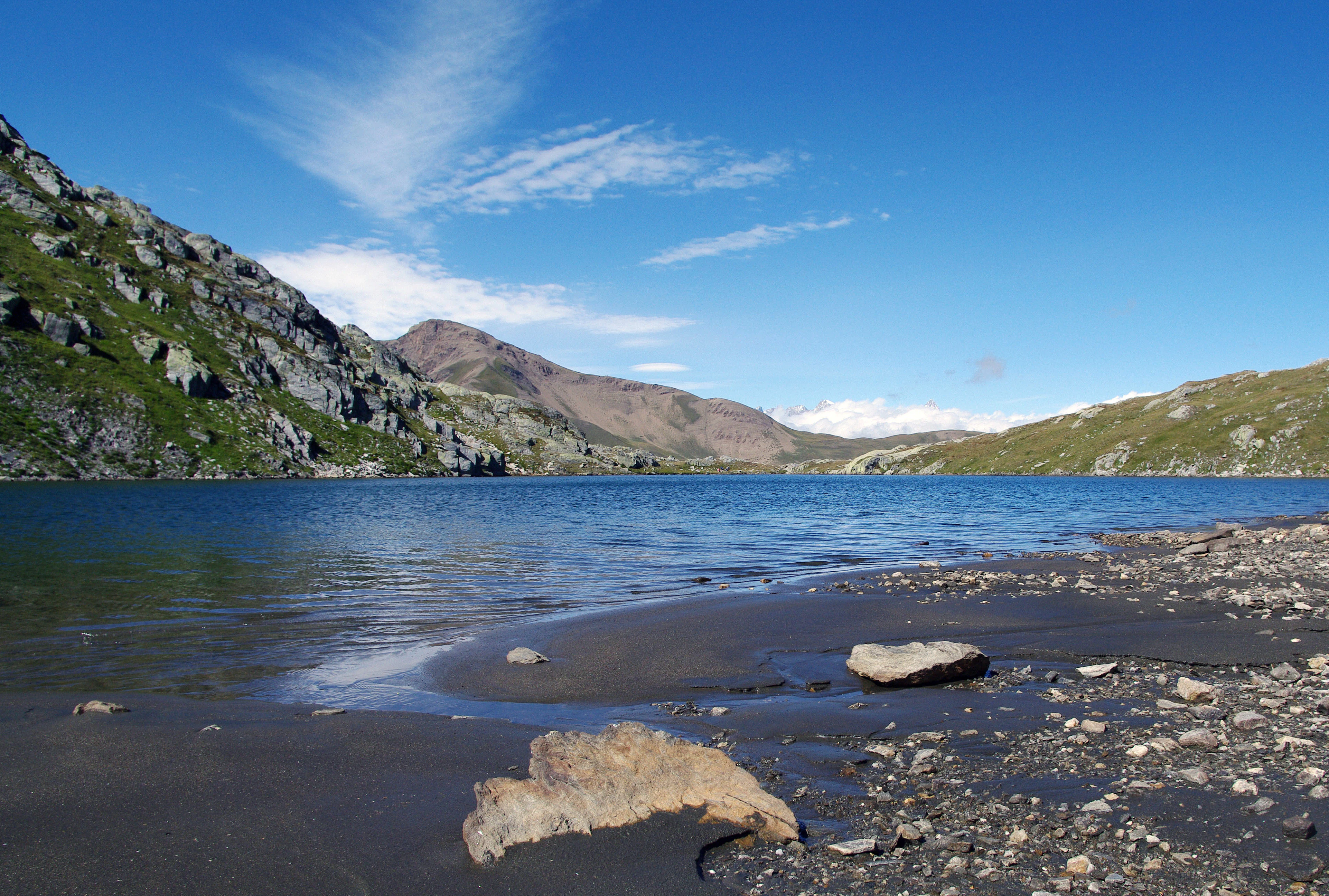 A calm, deep blue alpine lake with a dark, rocky shoreline in the foreground and steep mountain slopes rising in the background under a clear sky.