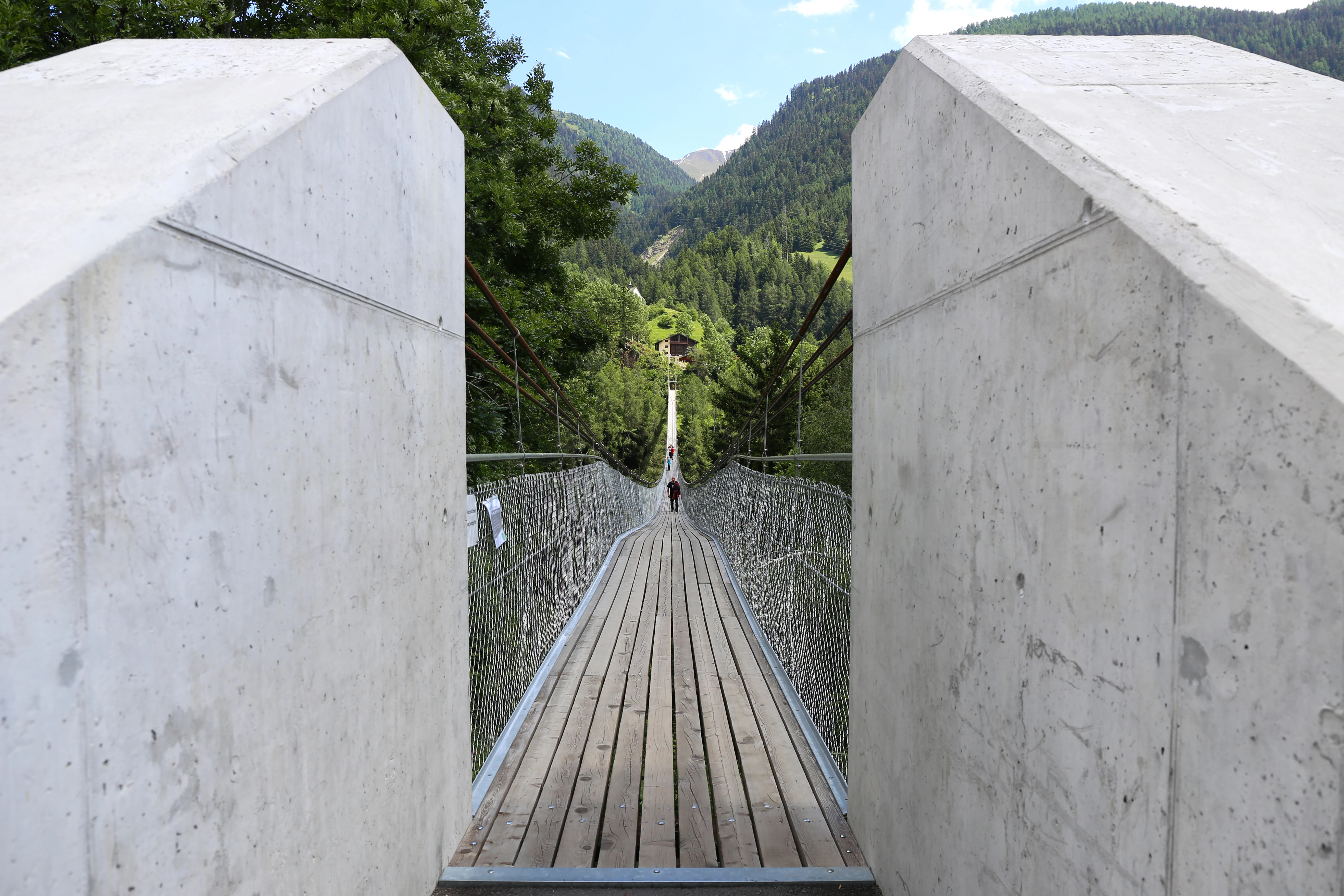 A view looking straight along the wooden deck of a suspension bridge, flanked by large, smooth concrete pillars at the entrance, with forested mountains visible in the distance.