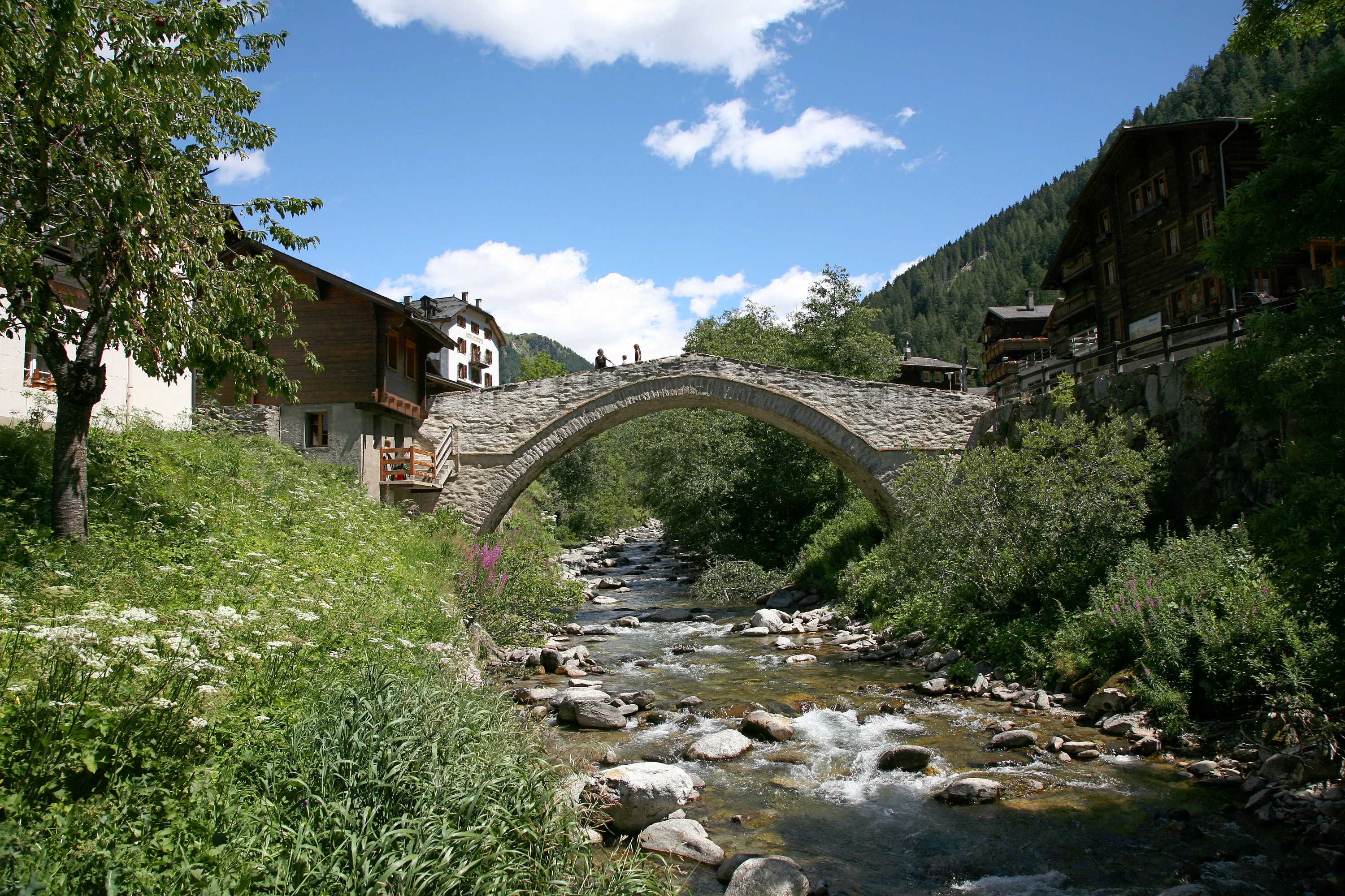 An arched stone bridge over a mountain stream in the Swiss village of Binn, surrounded by traditional wooden buildings and lush green vegetation.