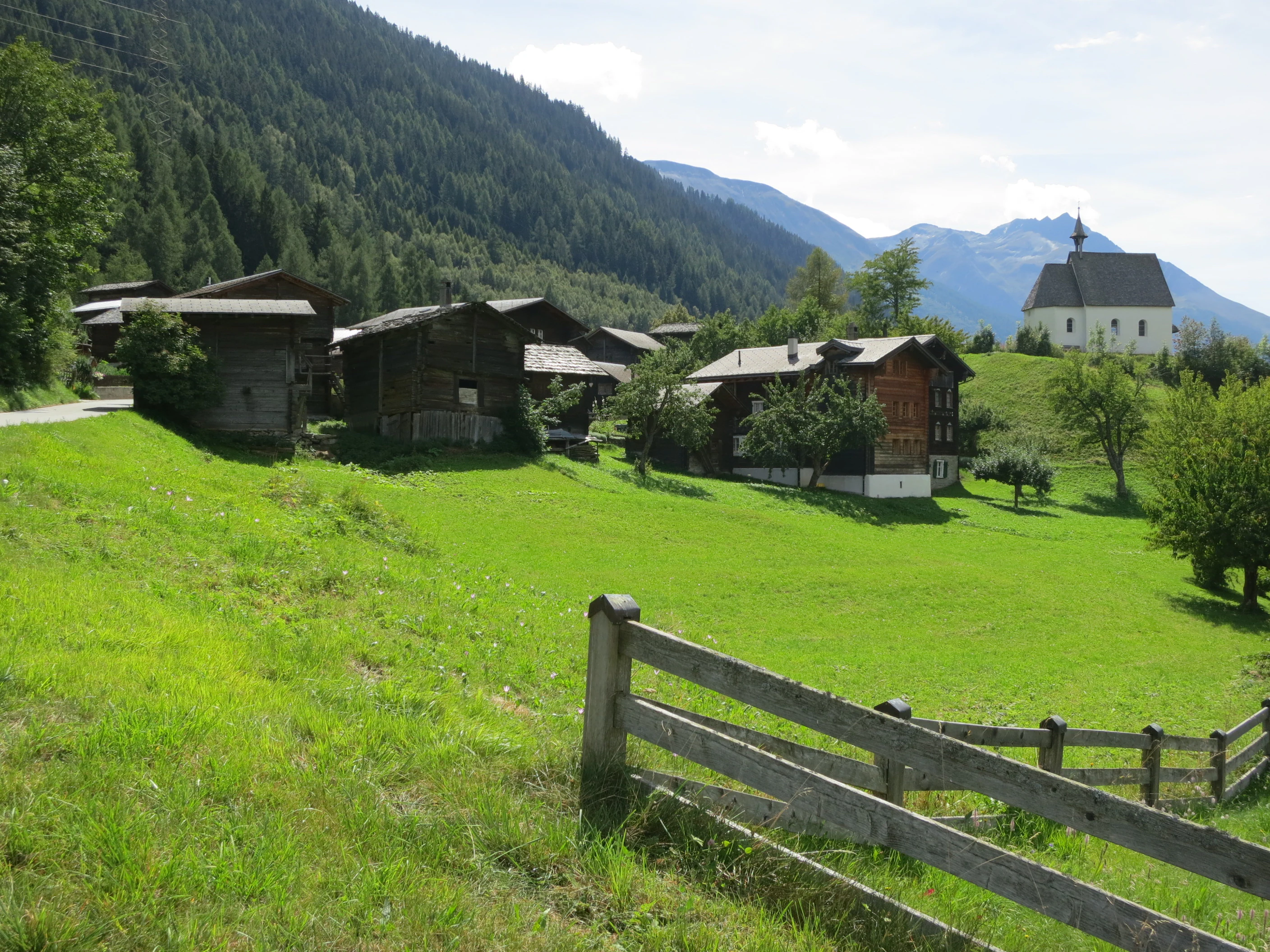 A rural Swiss mountain village with weathered wooden barns and a small white church, surrounded by green meadows, a wooden fence in the foreground, and steep forested mountain slopes in the background.