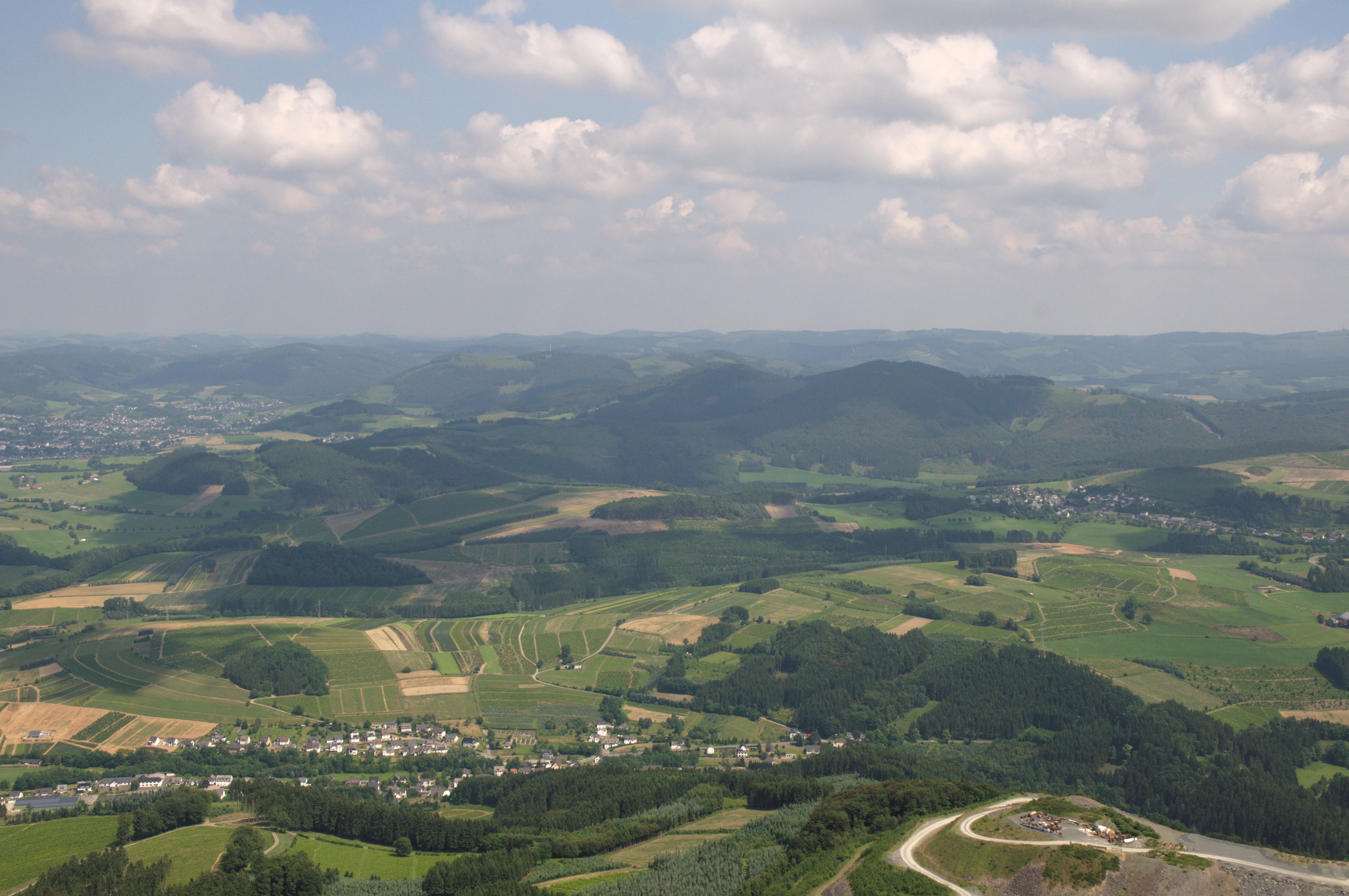 An aerial view showing a patchwork of green agricultural fields, forests, and small settlements nestled among rolling hills under a cloudy sky.