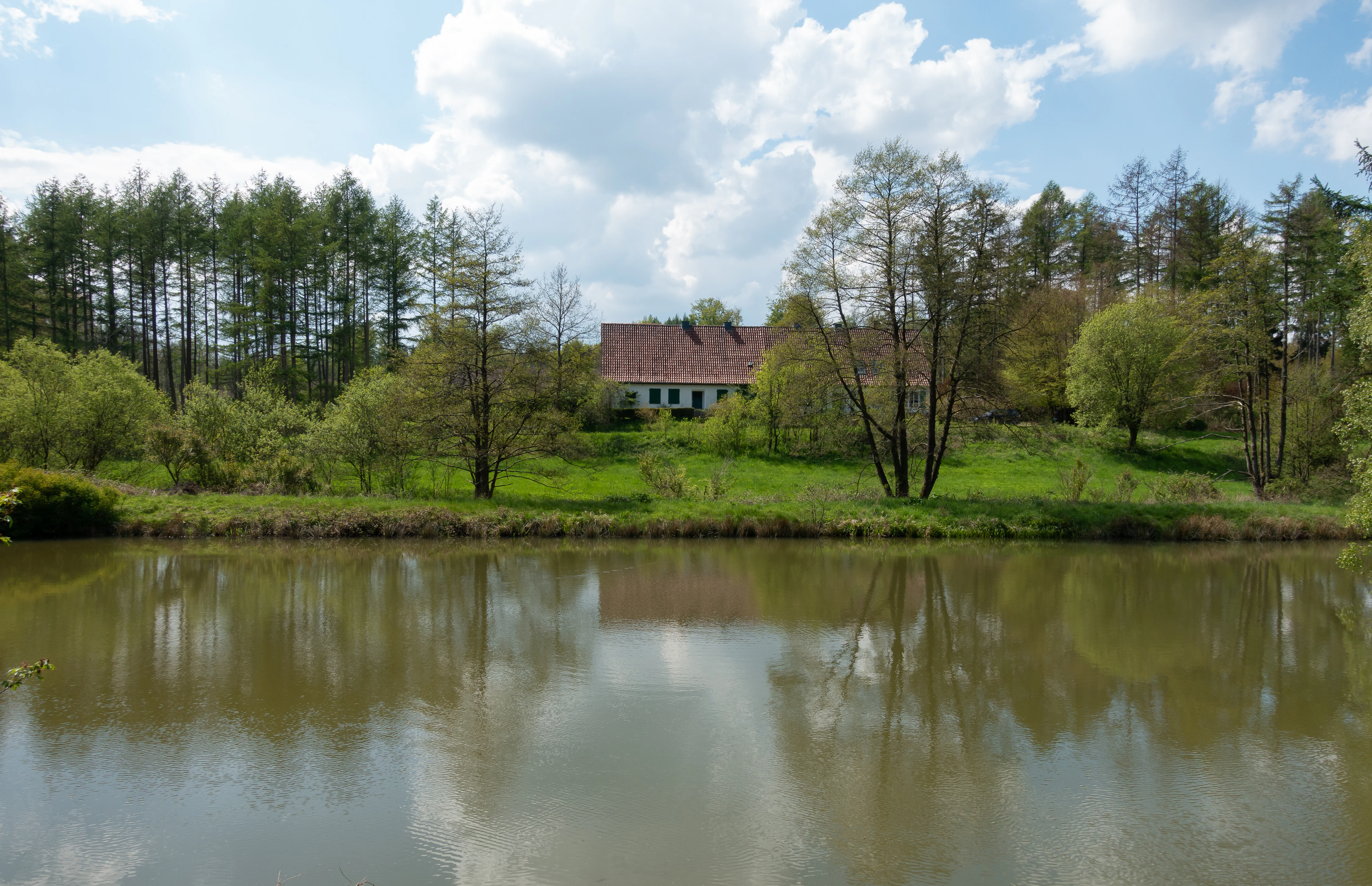 A still pond reflecting a historic building with a red tile roof and surrounding deciduous trees and forest under a partly cloudy sky.