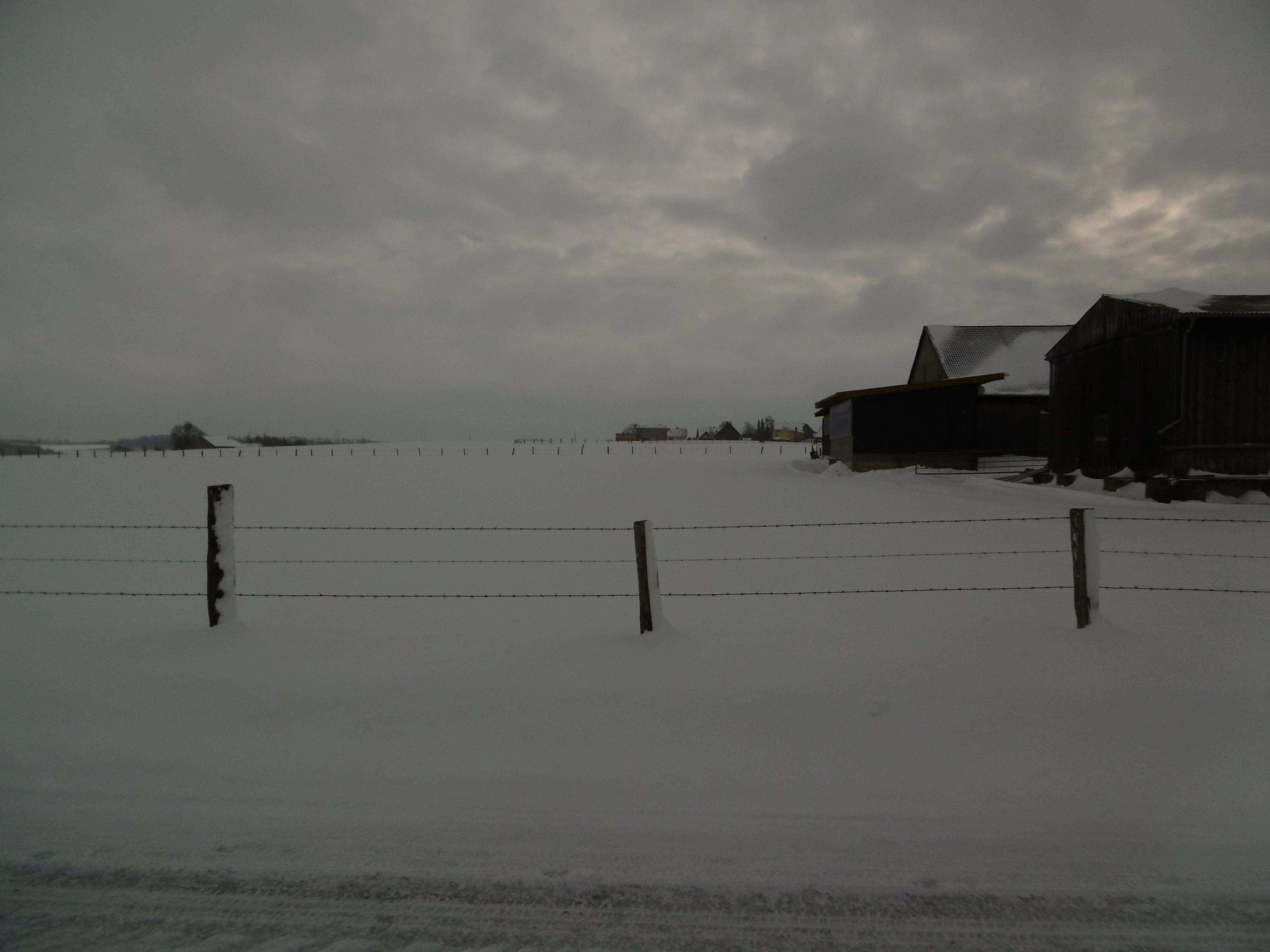 A wide shot of a snow-covered open field in Germany with a wooden fence in the foreground and farm buildings on the right under an overcast, grey sky.