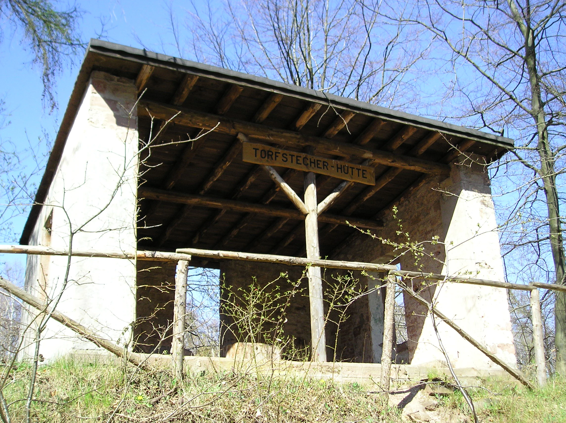 A small, open-walled stone shelter with a wooden roof, featuring a wooden sign reading Torfstecher-Hutte hanging from the central support beam.