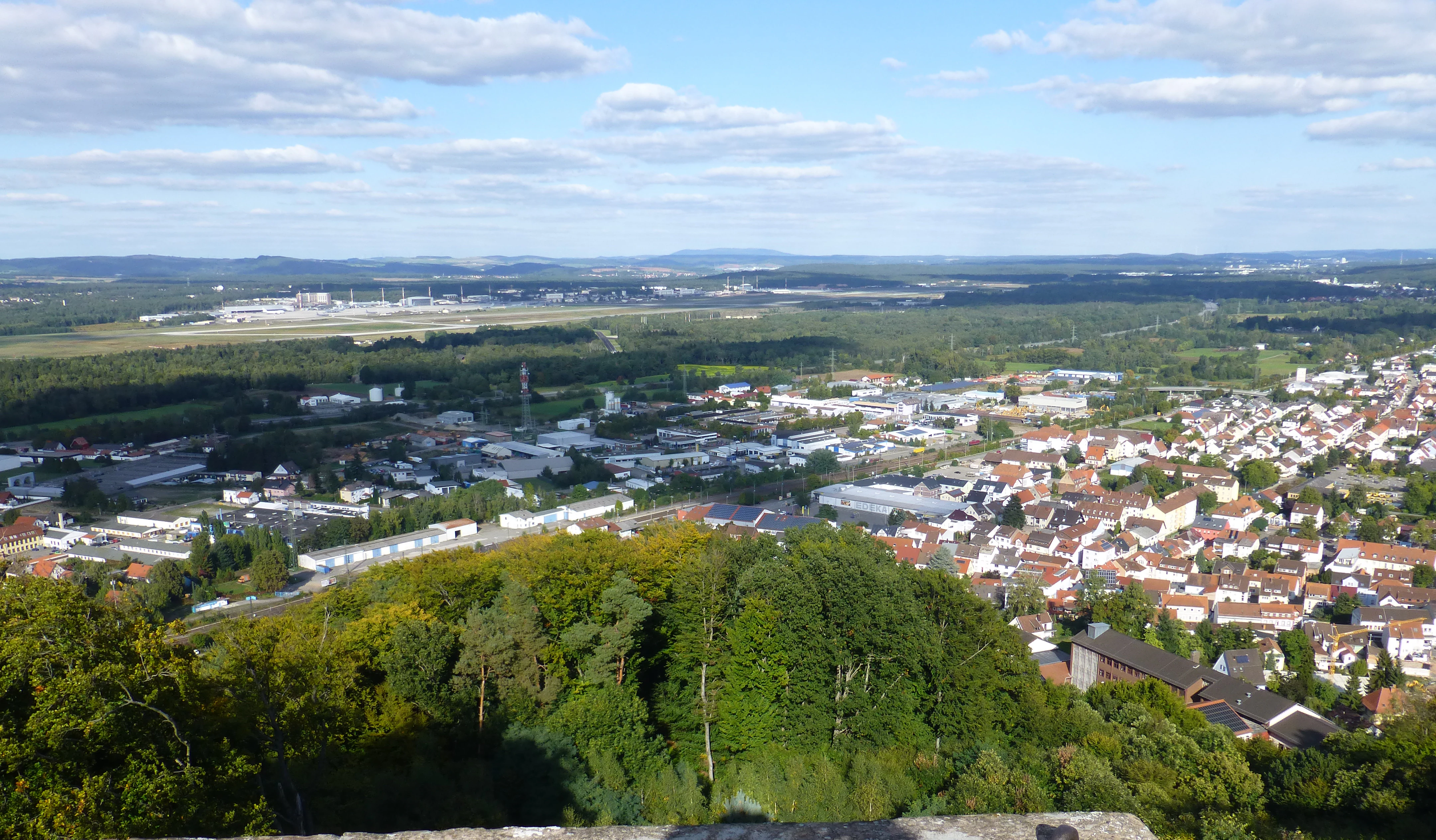 A high angle panoramic view from a tower looking down over a town with houses, forested hills, and a distant airfield under a blue sky with scattered clouds.