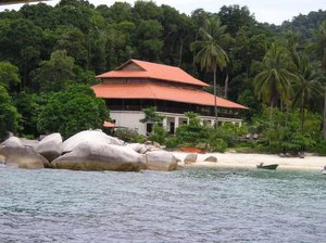 A view from the water showing a multi-tiered building with a red tile roof set against a backdrop of dense green forest, with large granite boulders and a sandy beach in the foreground.