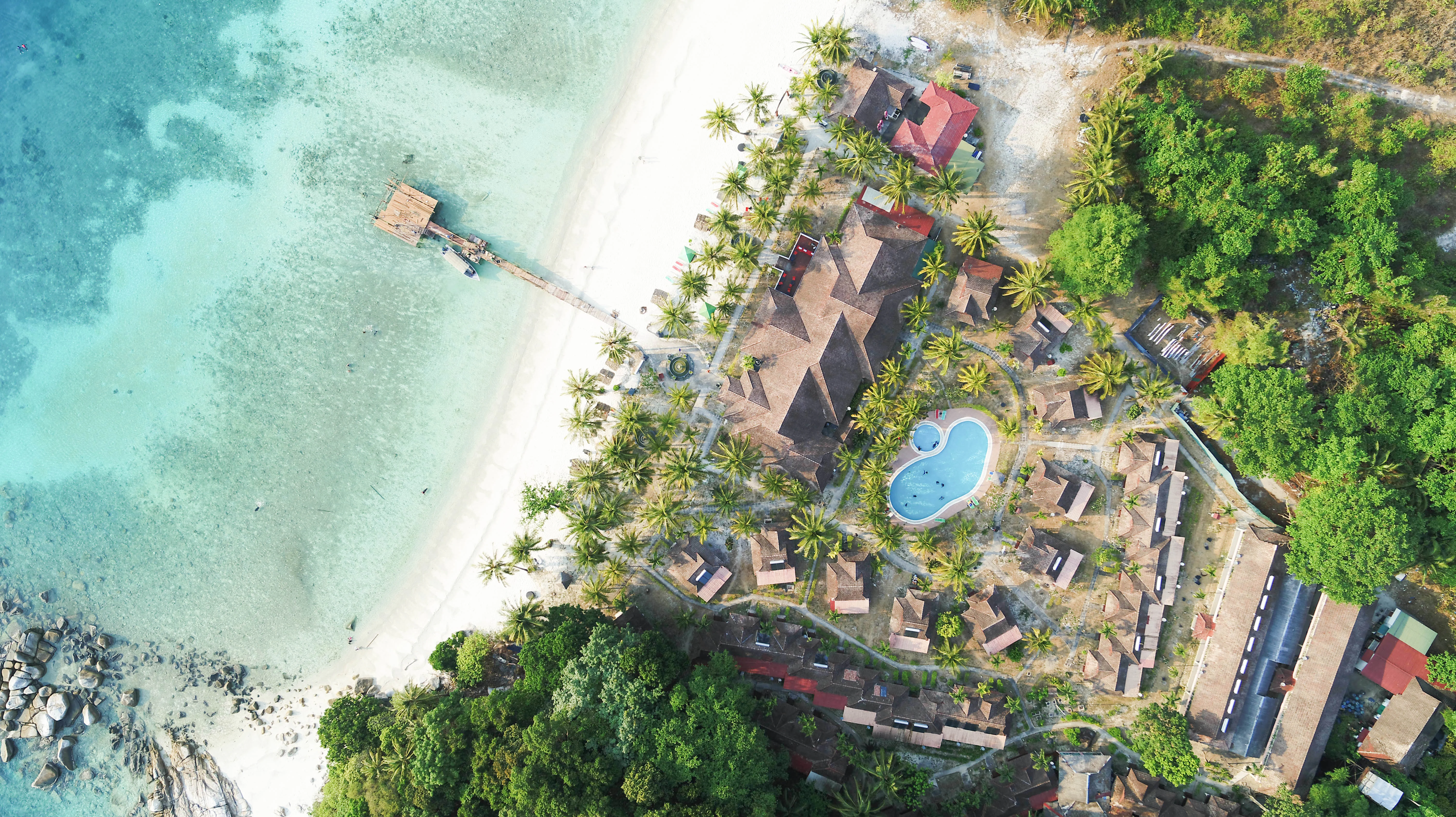 An aerial view showing a beach resort on Lang Tengah Island, with white sand beach, turquoise water, a wooden pier, and resort buildings nestled in tropical vegetation.