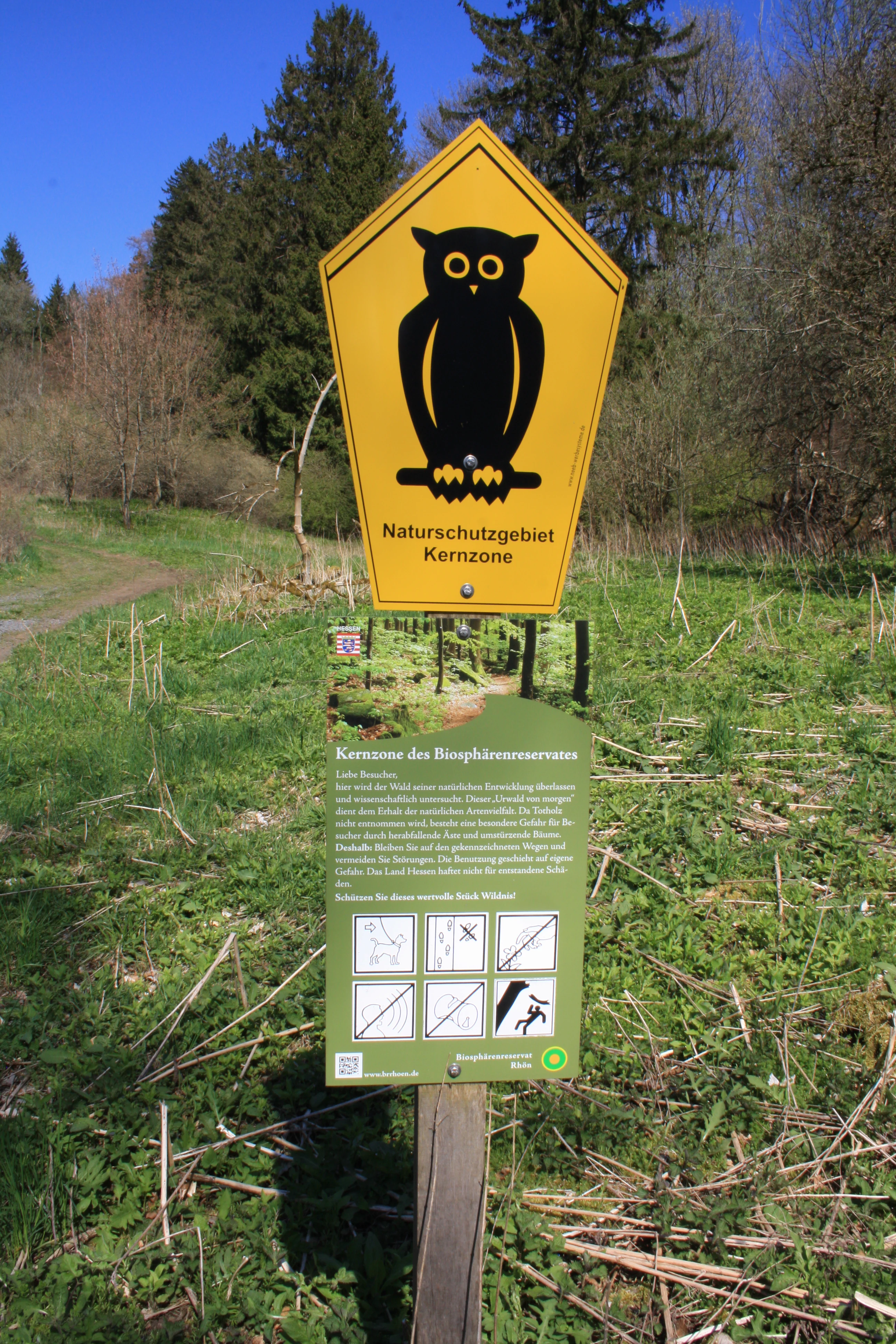 A yellow pentagonal nature reserve sign featuring a black owl icon, mounted above a secondary green information board with various pictograms and text, set in a grassy meadow with trees in the background.