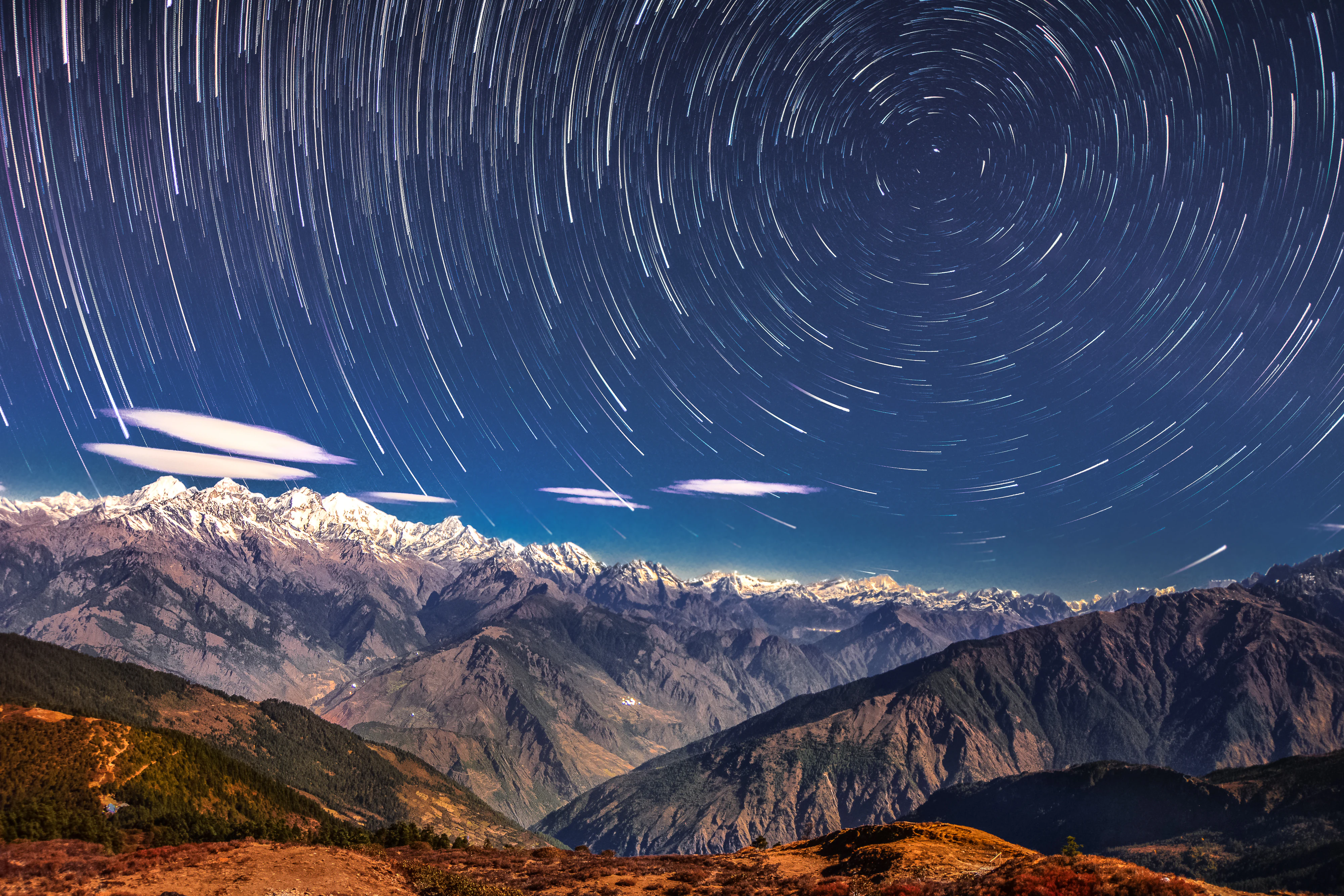 Circular star trails over mountain peaks under a dark night sky with scattered clouds