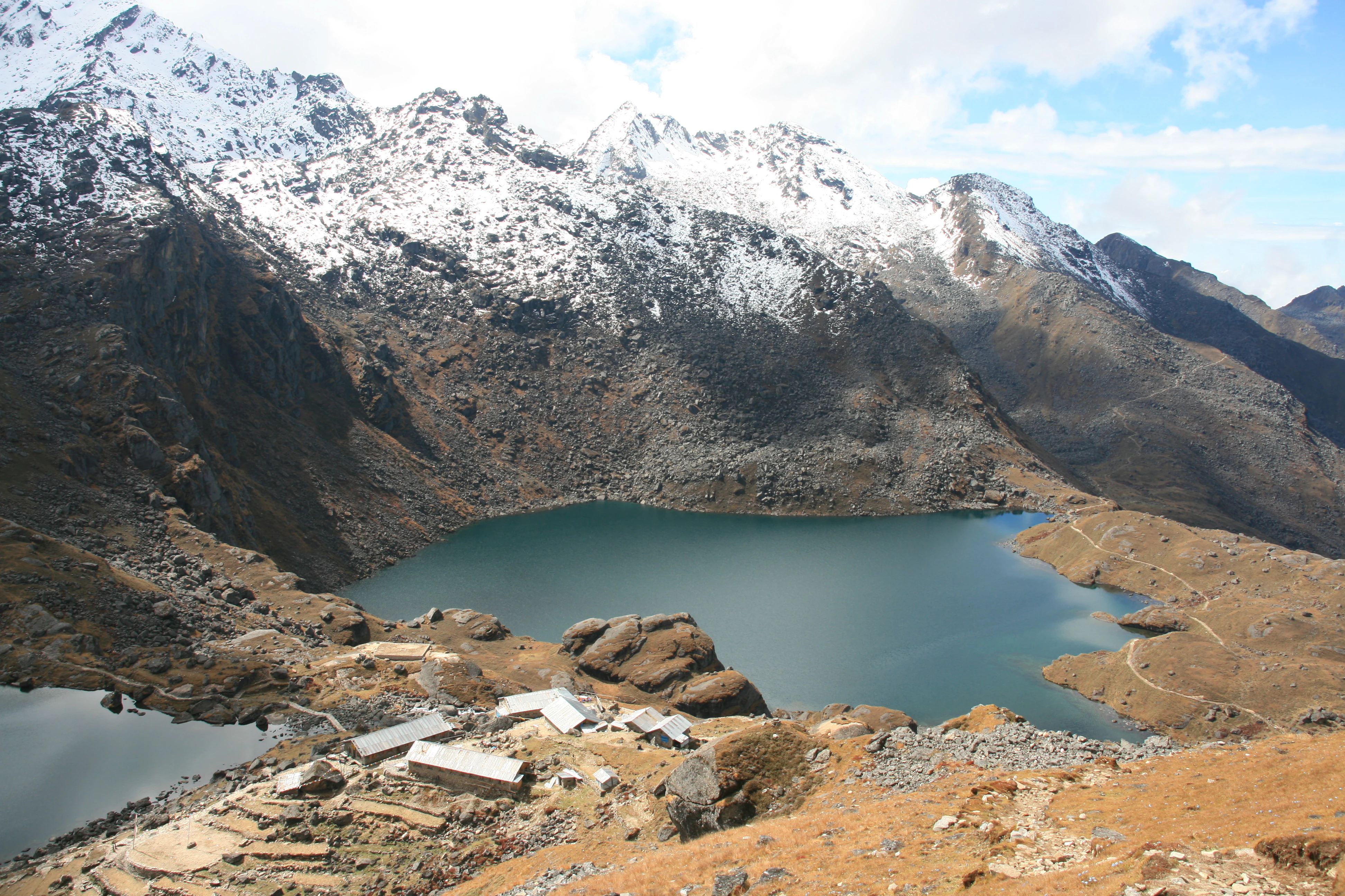 Snow-capped mountain peaks overlook a turquoise lake with rocky terrain and small structures nearby