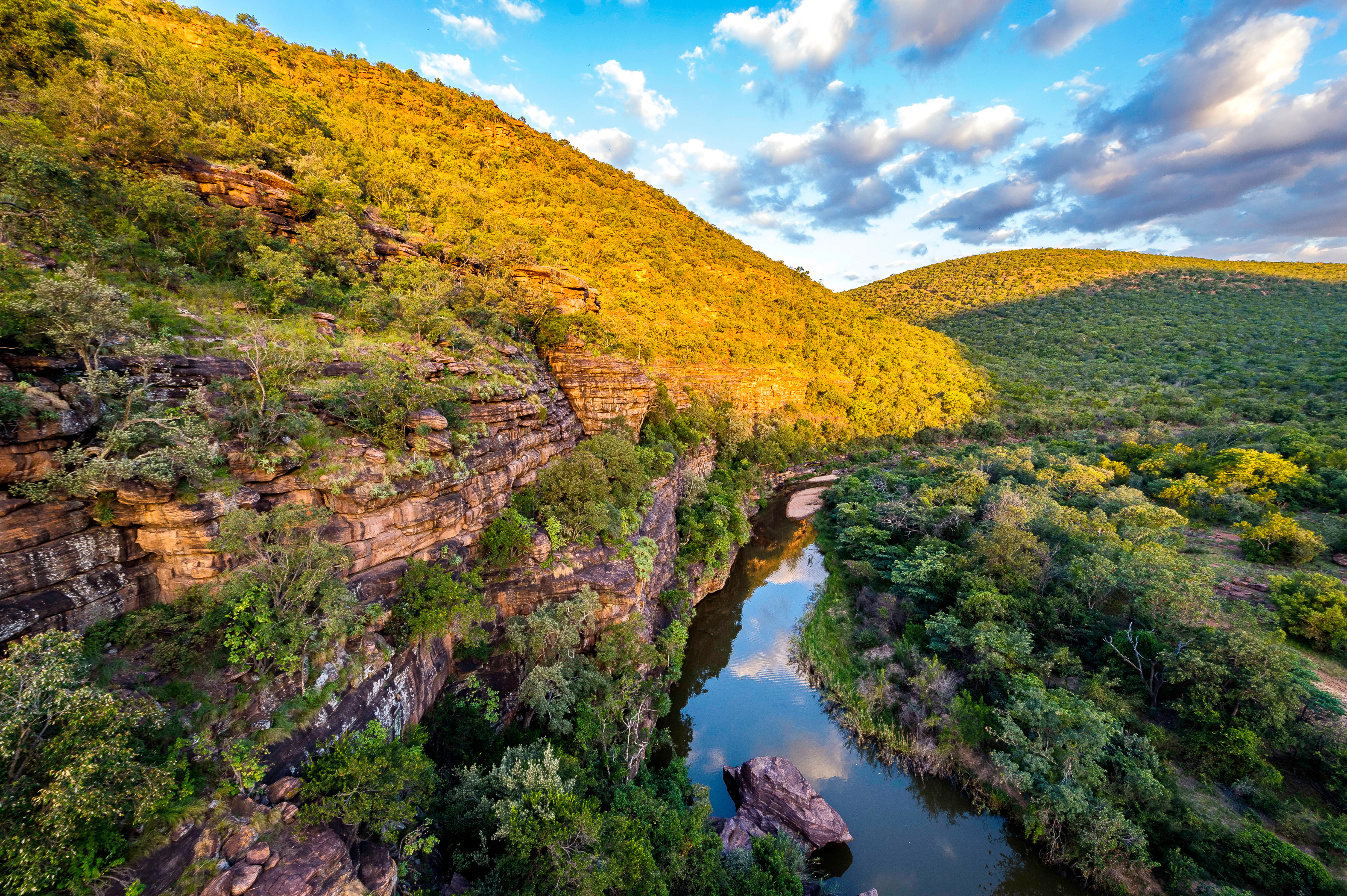 A high-angle view of a calm, reflective river flowing through a deep valley between steep, tree-covered rocky hills under a partially cloudy blue sky.