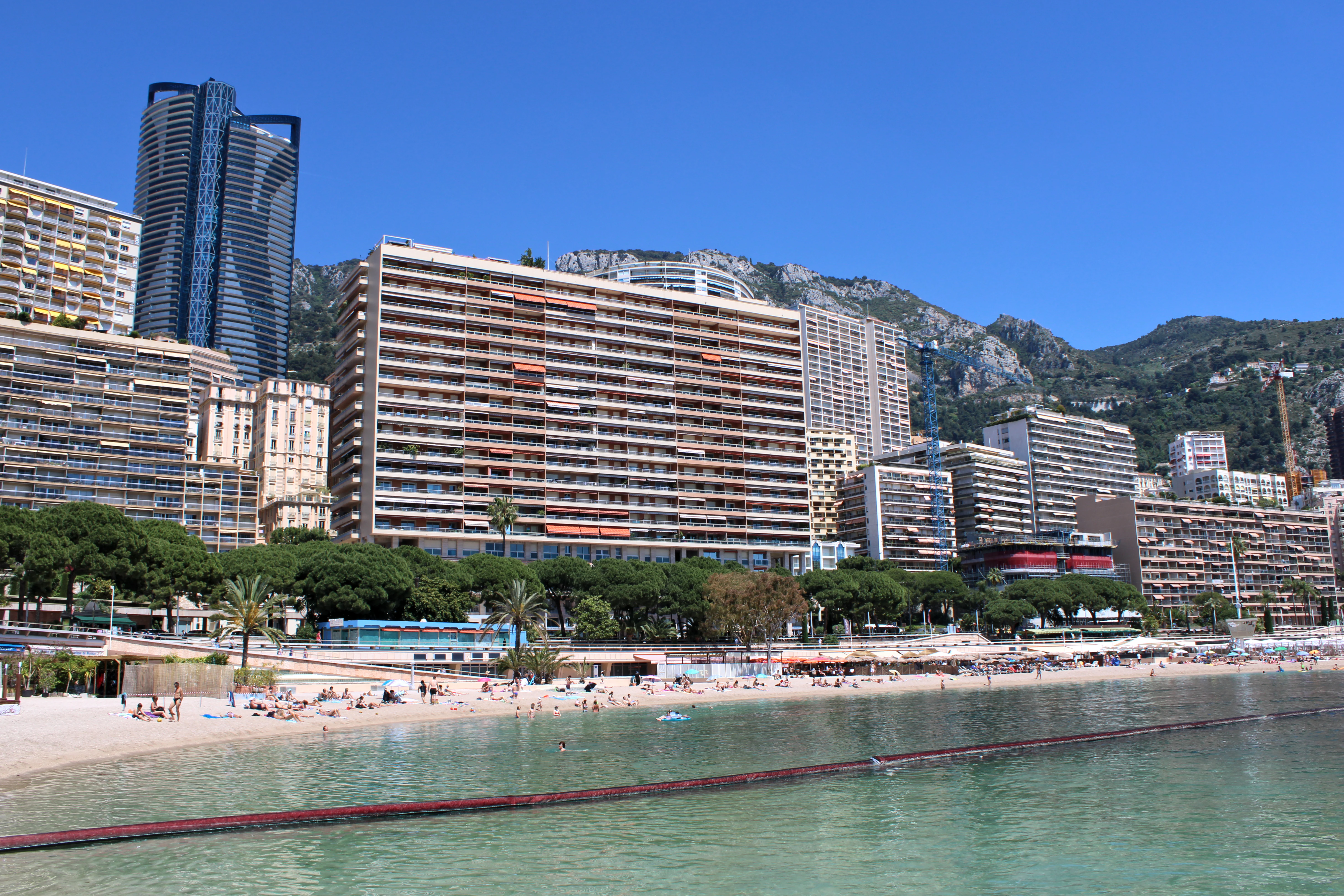 A wide angle view of the Larvotto beach in Monaco, featuring a sandy beach, clear water, and a dense backdrop of residential high rise buildings against a clear blue sky.