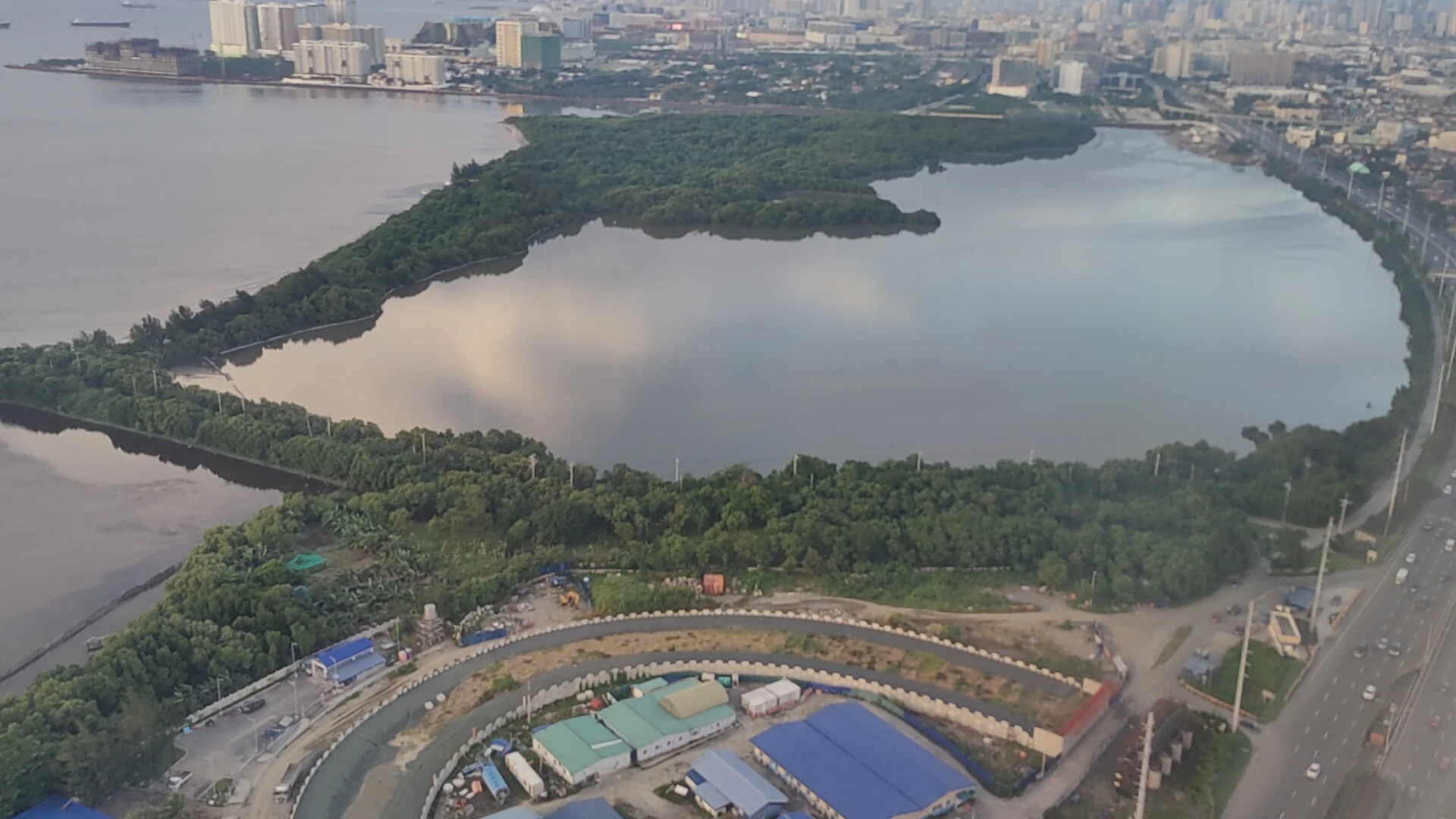 Una toma aérea que muestra los manglares verdes y el agua oscura del parque de humedales, bordeado por desarrollo urbano y una autopista.