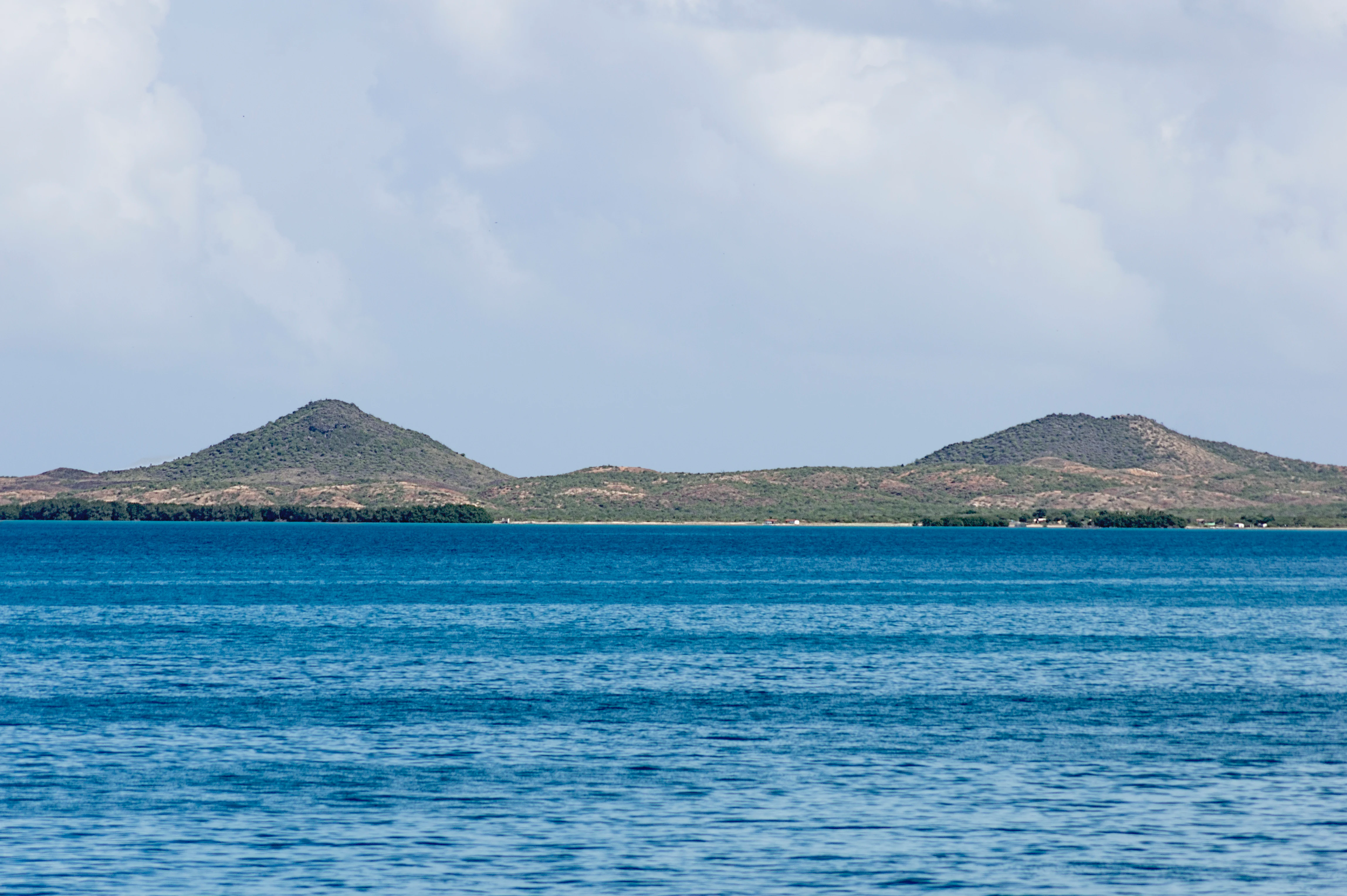 Two prominent, rounded twin hills rise above a calm blue coastal landscape under a cloudy sky.