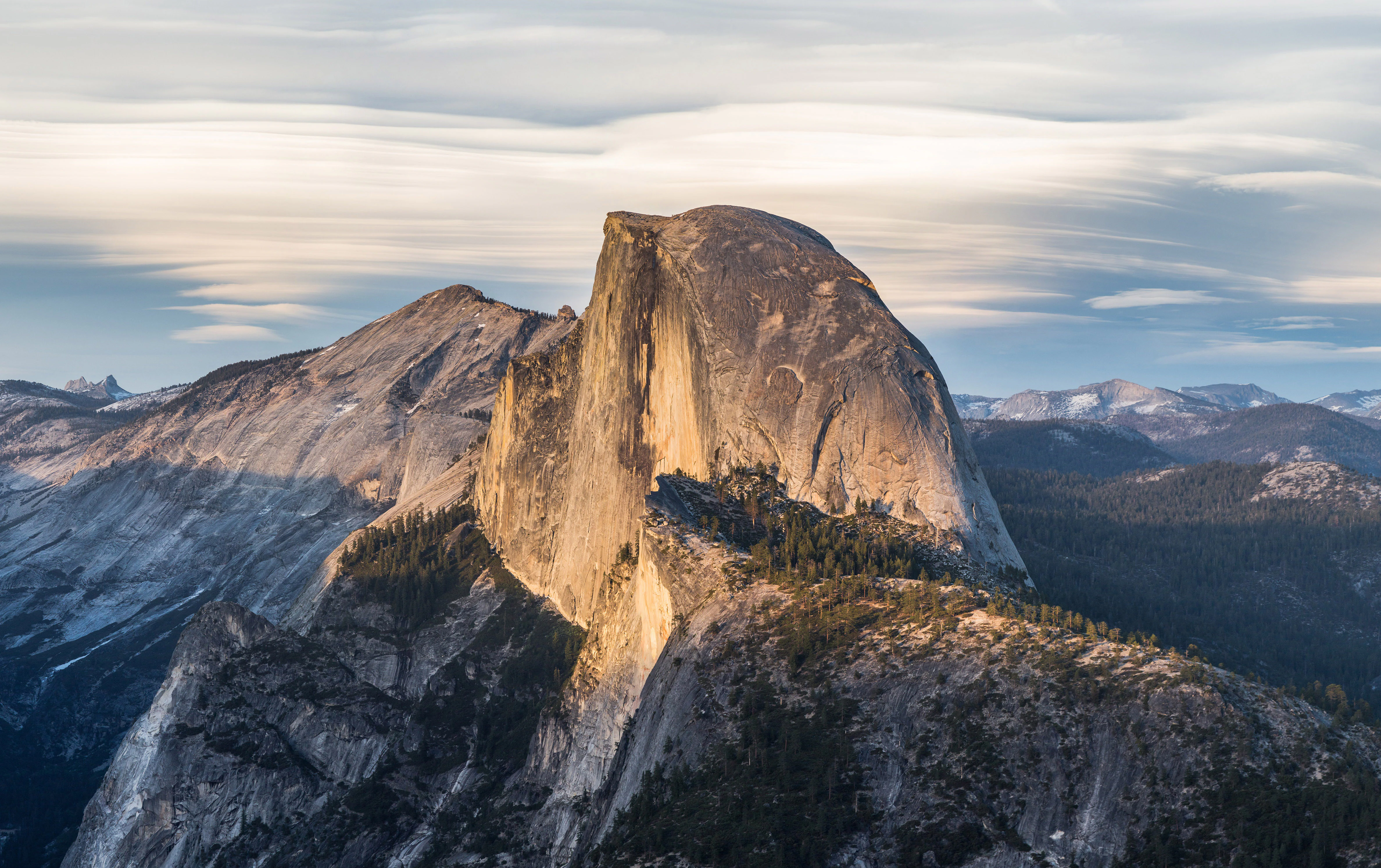 A large granite dome mountain illuminated by warm sunlight with layered clouds above and forested slopes below in a mountainous landscape