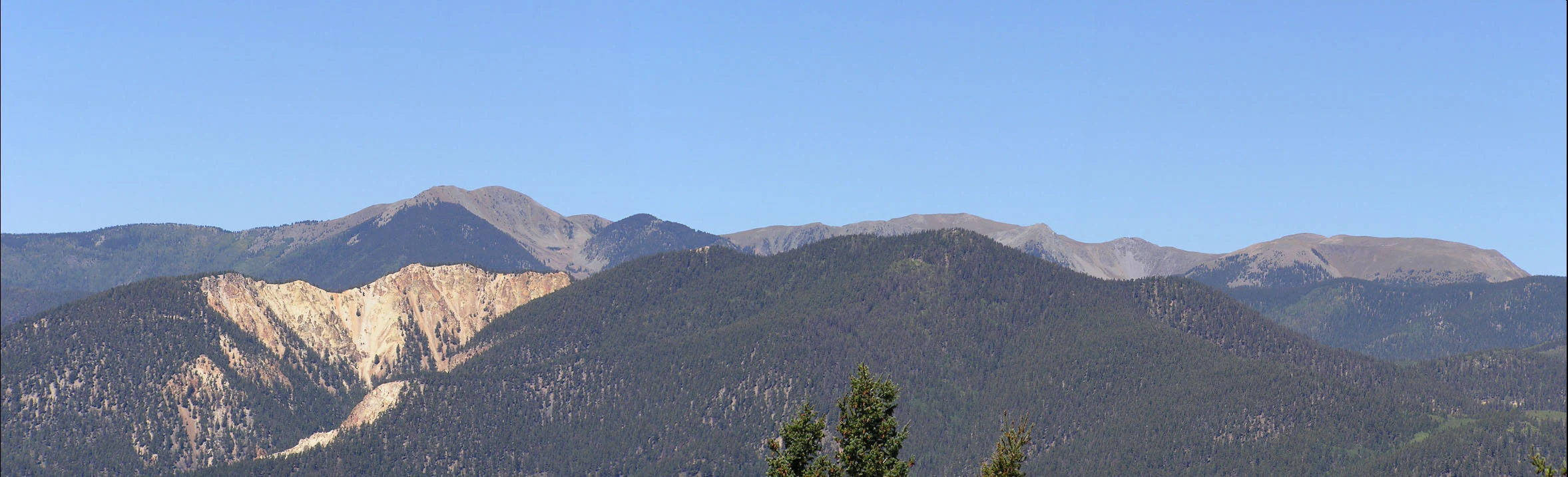 A wide, panoramic view of a mountain range under a clear blue sky, featuring dark, pine-covered slopes and exposed rocky summits.