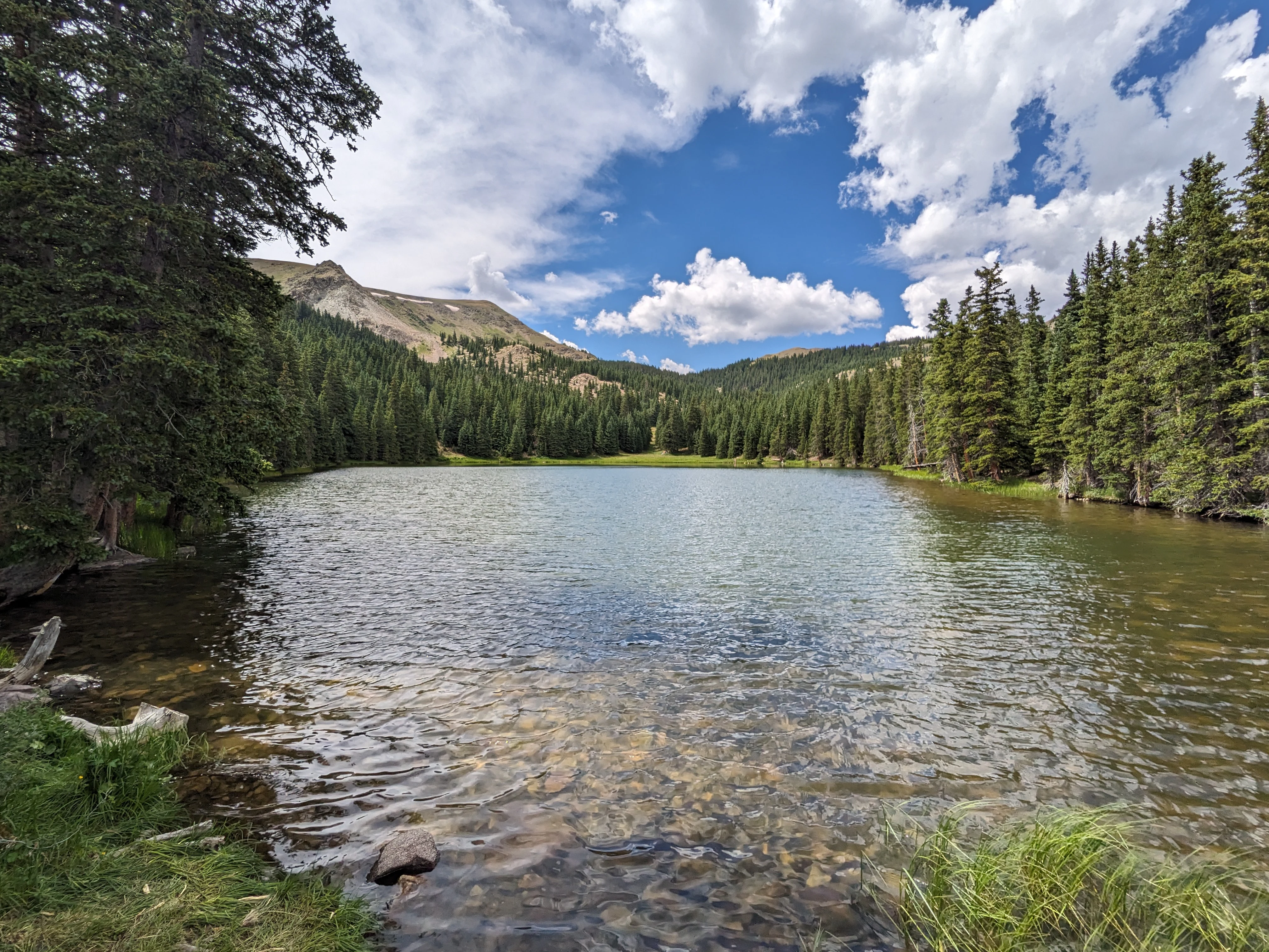 A clear alpine lake surrounded by dense evergreen forests and mountain slopes under a partly cloudy sky.