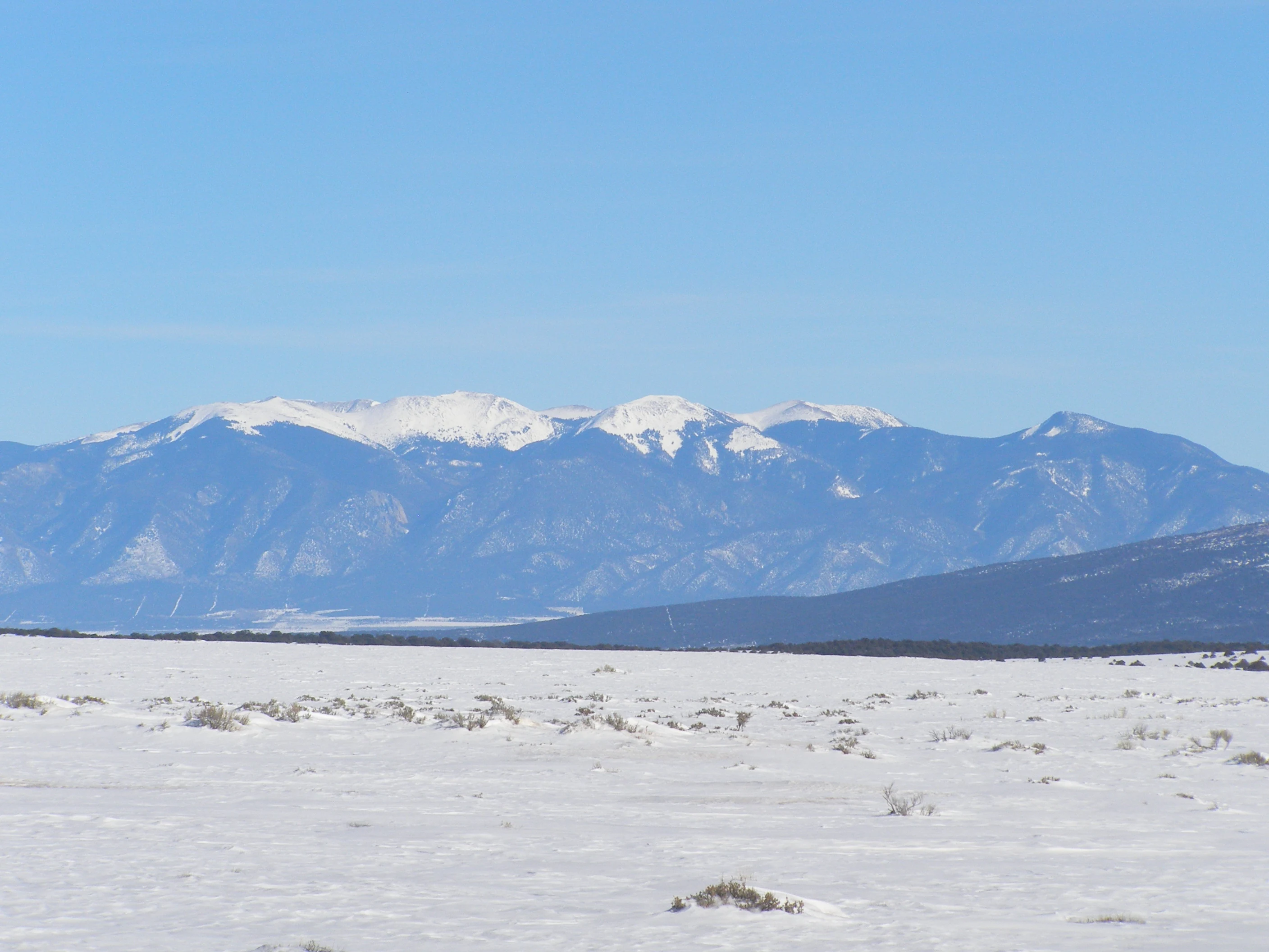 A distant, snow-capped mountain range rises above a flat, snow-covered plain under a clear blue sky.