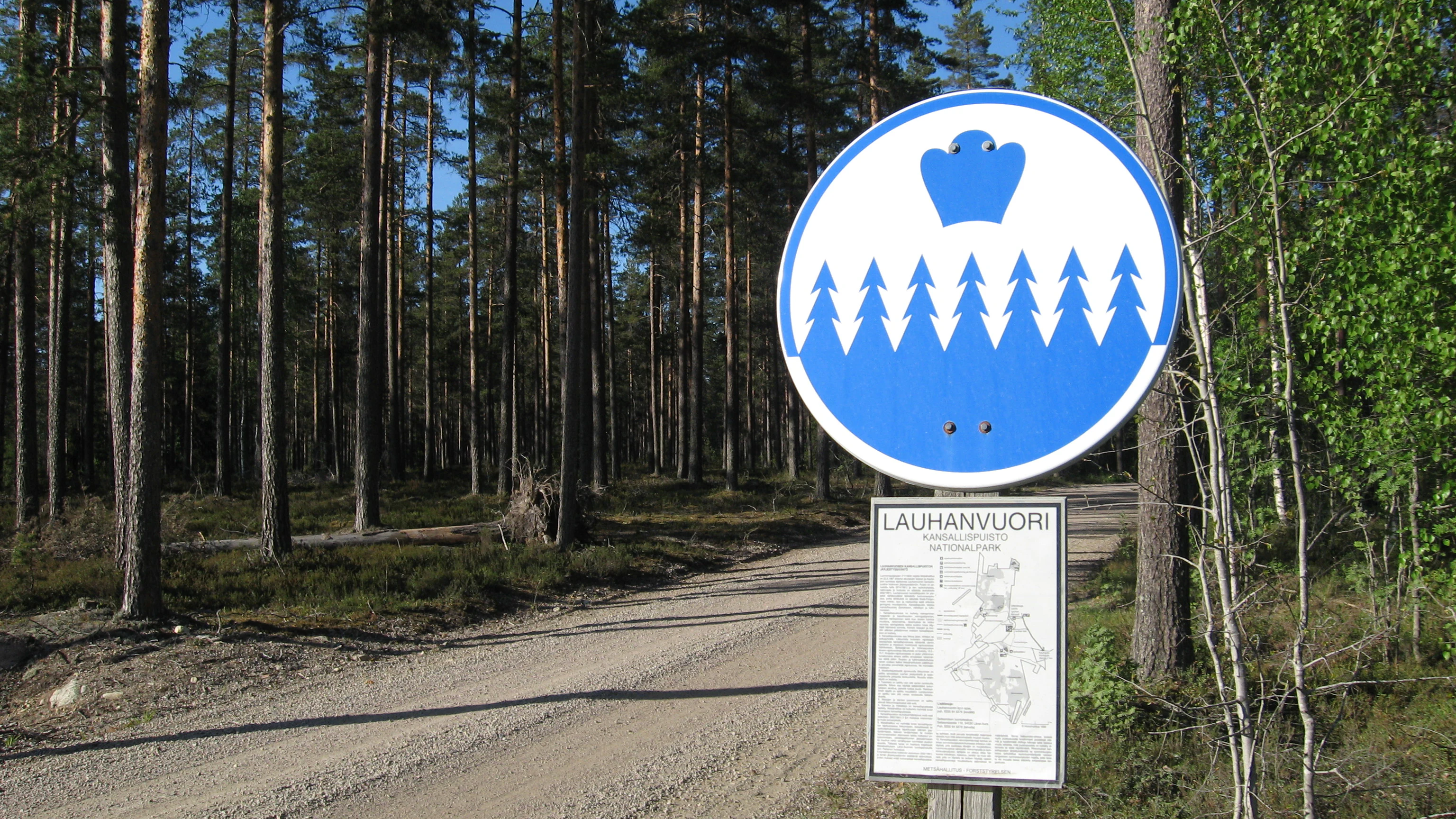 Circular blue and white park boundary sign with crown and pine tree symbols, mounted above an informational sign reading LAUHANVUORI, on a dirt road surrounded by pine forest