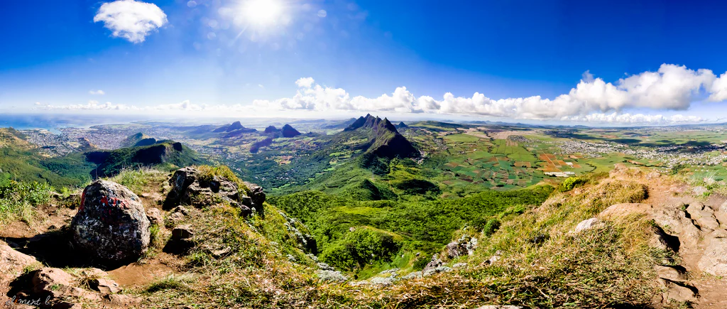 Una vista panorámica de gran angular tomada desde un pico rocoso de montaña que mira hacia un valle frondoso y verde y colinas onduladas bajo un cielo azul brillante con nubes dispersas.