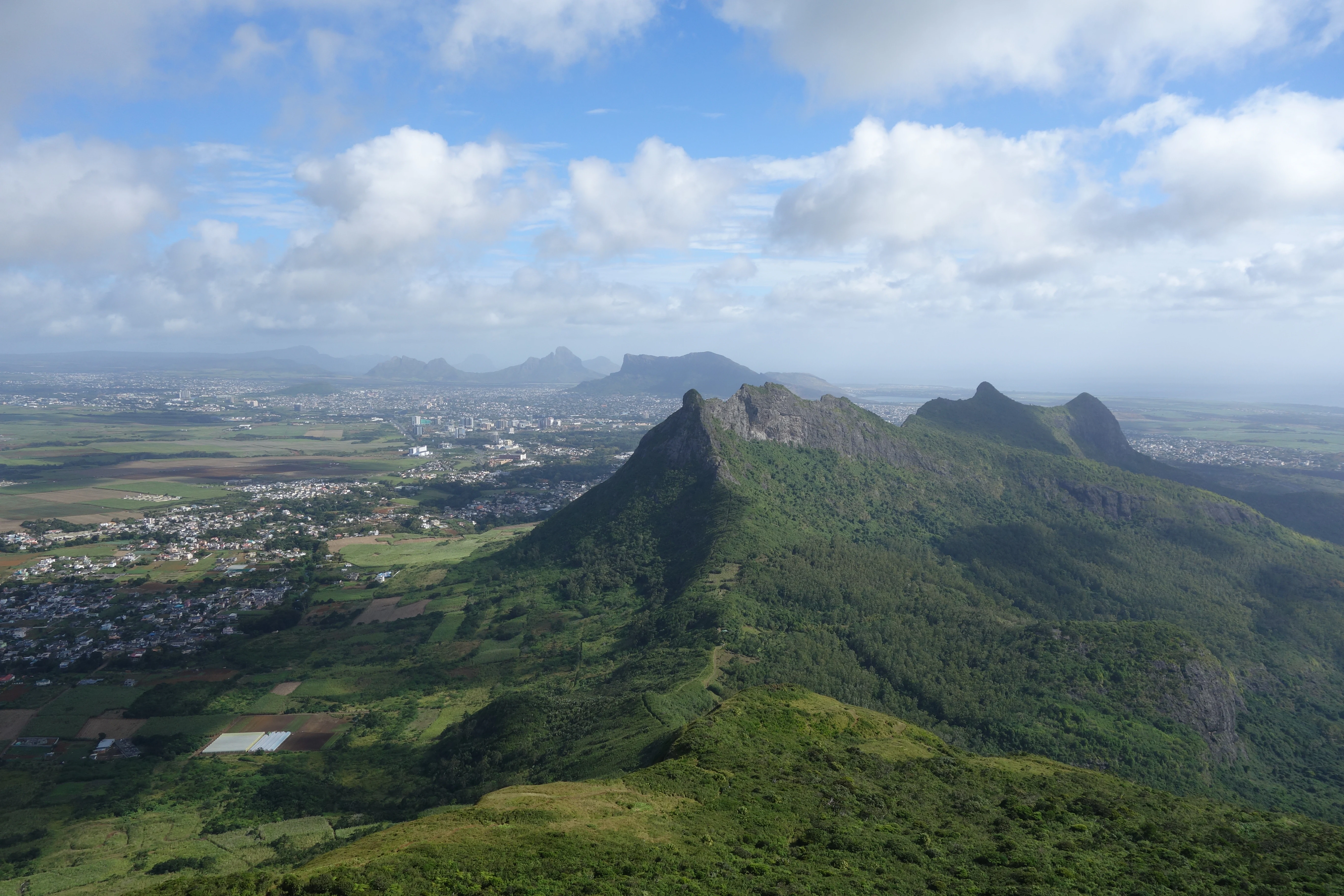 Vista de gran altitud desde la cima de una cadena montañosa, con vistas a colinas verdes boscosas, campos agrícolas y un pueblo lejano, bajo un cielo azul con nubes dispersas.