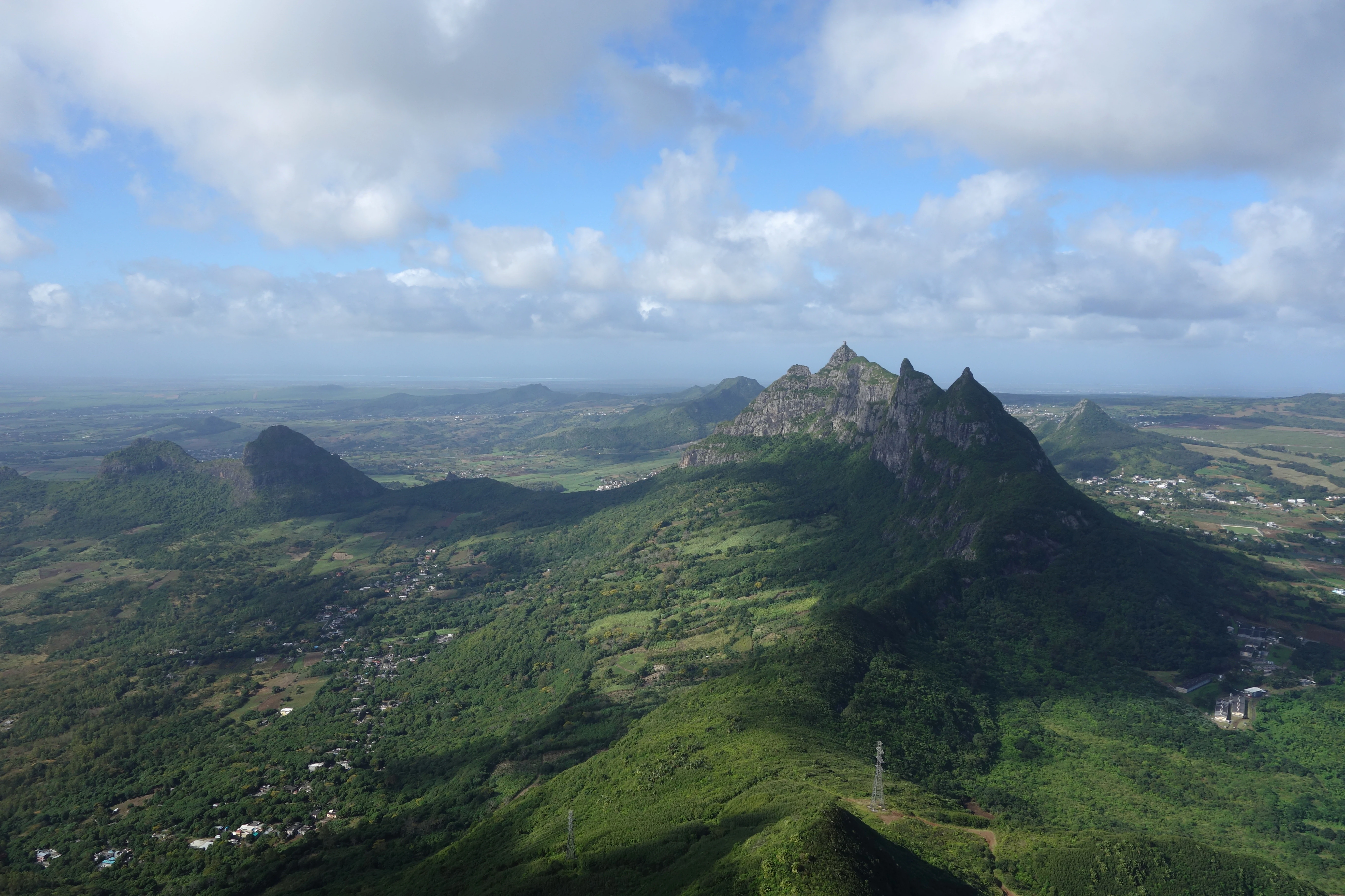 Una amplia vista aérea del paisaje tomada desde un pico montañoso elevado, con vistas a un exuberante valle tropical verde hacia un pico montañoso escarpado y prominente en la distancia, bajo un cielo parcialmente nublado.