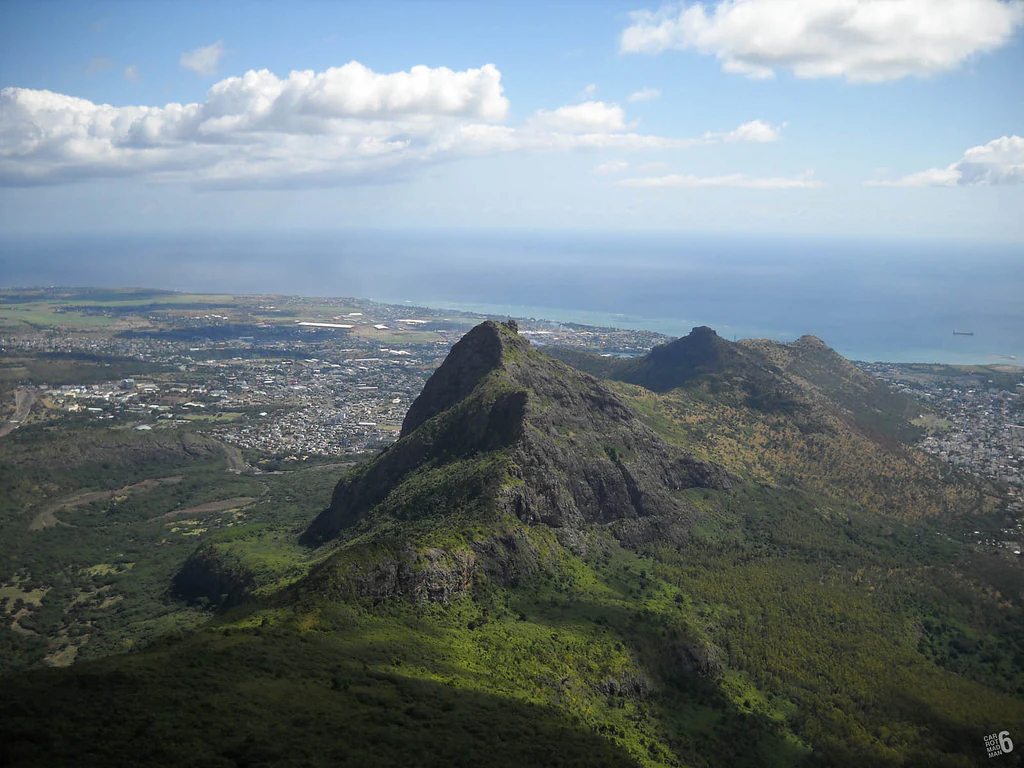 Vista aérea en ángulo alto desde la cima de una montaña sobre una cresta rocosa y verde, con una ciudad extensa y el océano visibles al fondo bajo un cielo azul parcialmente nublado.