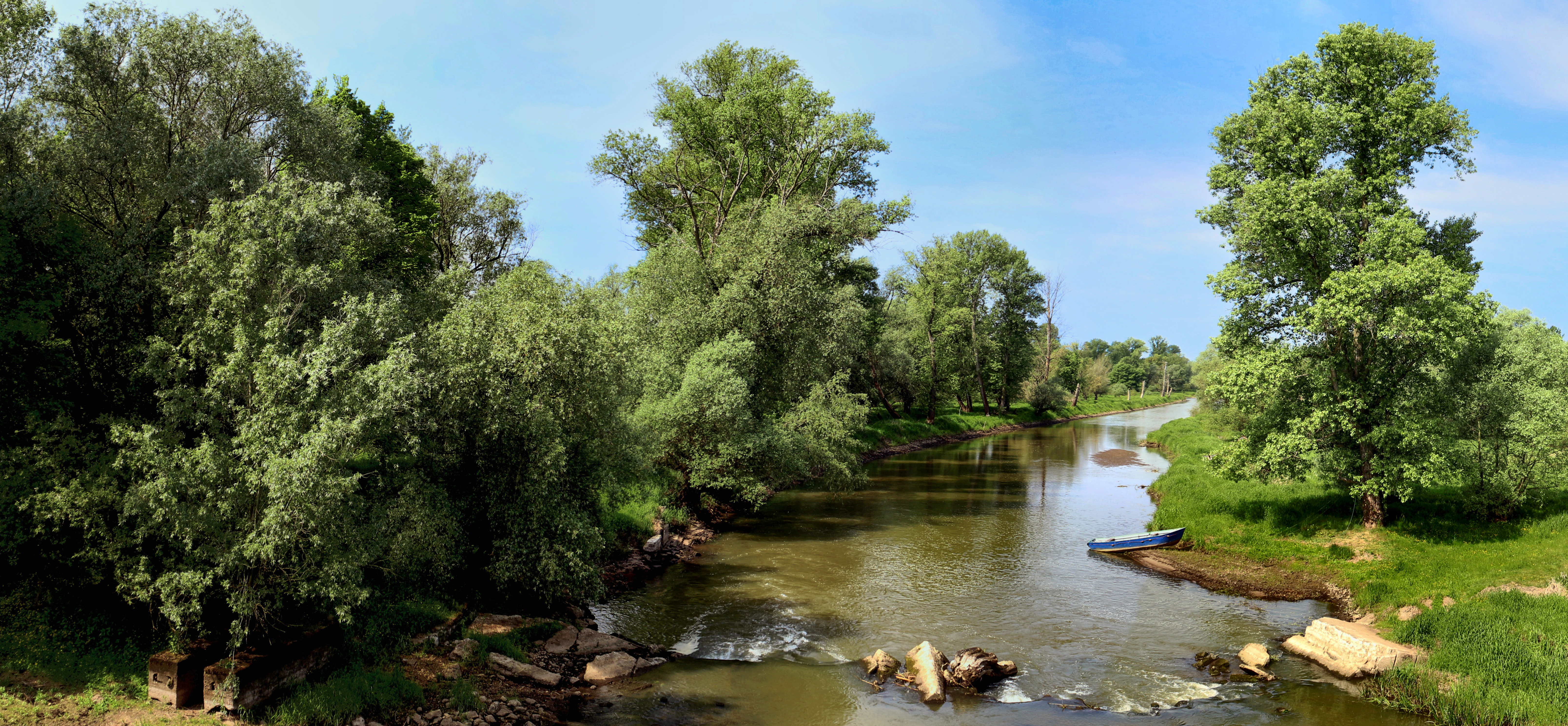 A wide panoramic shot of a river flowing through lush green trees, with a small blue boat moored on the bank.